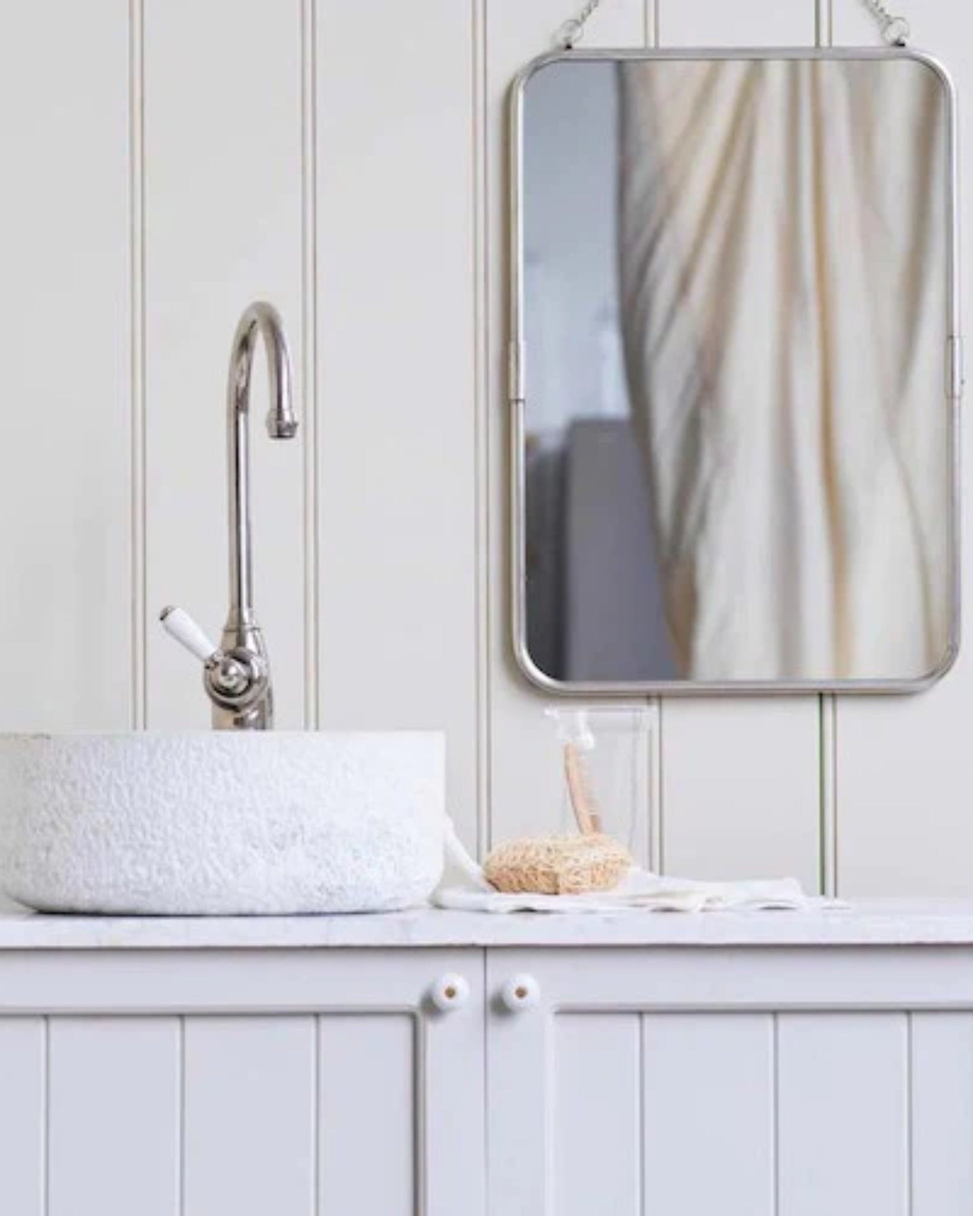 A minimalist bathroom features a white stone sink paired with a sleek metal faucet resting on a white cabinet. Above it, a rectangular mirror with a metal frame reflects part of a curtain, while the FIREHOUSE CERAMIC KNOB by Society Ink adds subtle elegance. A soap dispenser and brush sit on the countertop, completing the serene setup.