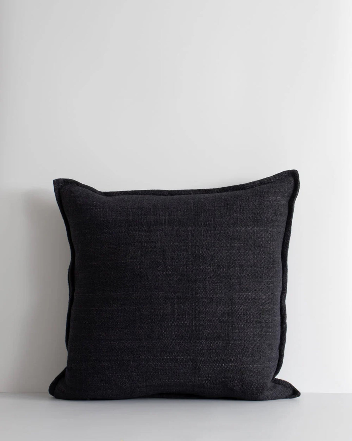 The FLAXMILL HANDWOVEN LINEN CUSHION by Baya, in a cloudburst shade, is positioned against a light gray backdrop. This dark gray, square cushion showcases raw linen fibers and a minimalist design, highlighted by subtle stitching along its edges.