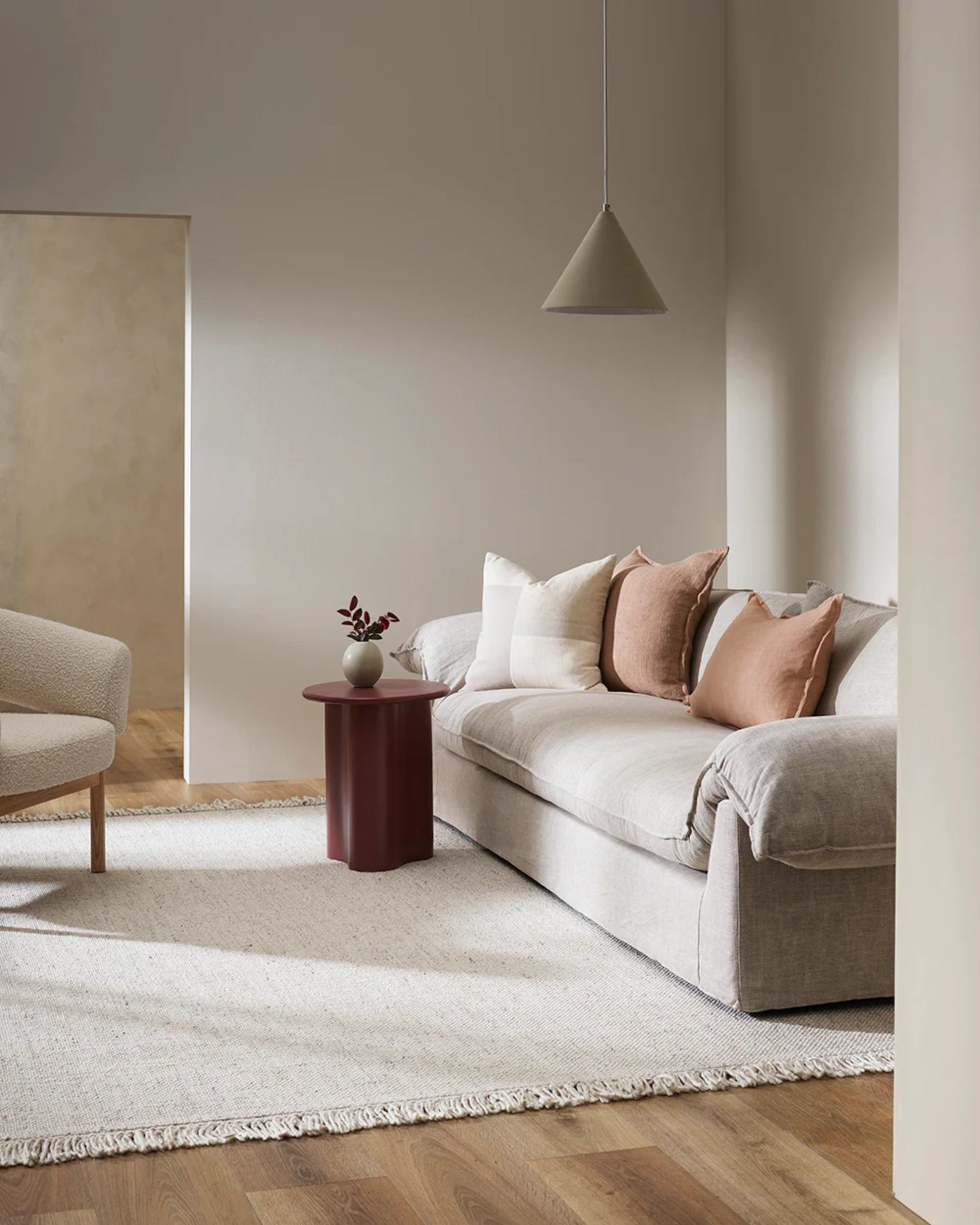 A minimalist living room features a beige sofa adorned with Baya's FLAXMILL HANDWOVEN LINEN CUSHION, complemented by a round maroon side table holding a small vase. A light beige armchair and neutral rug cover the wooden floor, while a pendant lamp hangs above. Natural light fills the space beautifully.