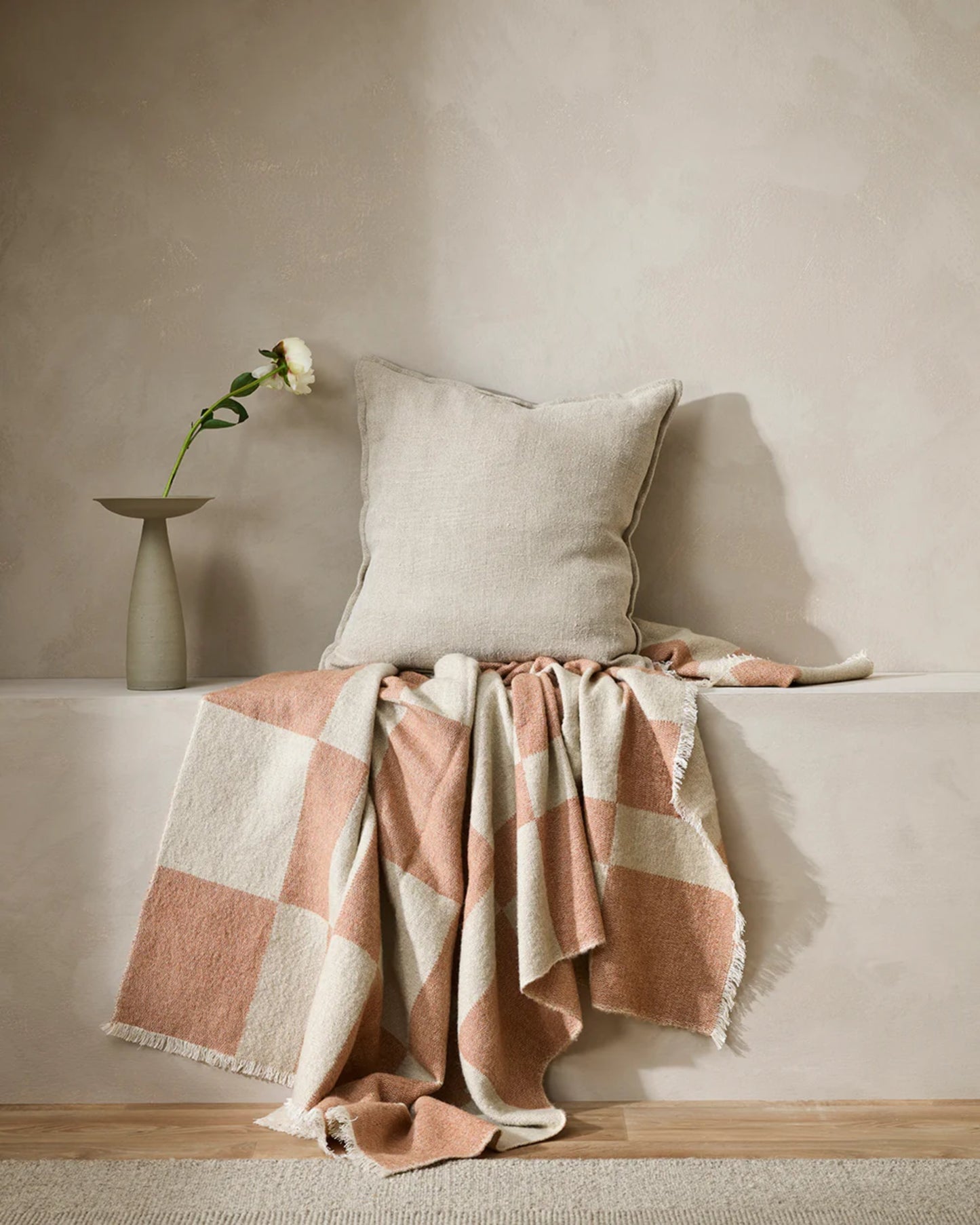 A cozy setup features the FLAXMILL HANDWOVEN LINEN CUSHION by Baya and a pink and white checkered throw blanket with a heavyweight weave on a light-colored bench. A single white flower in a minimalist vase is placed beside them against a textured wall, enhancing the cloudburst tone of the setting.