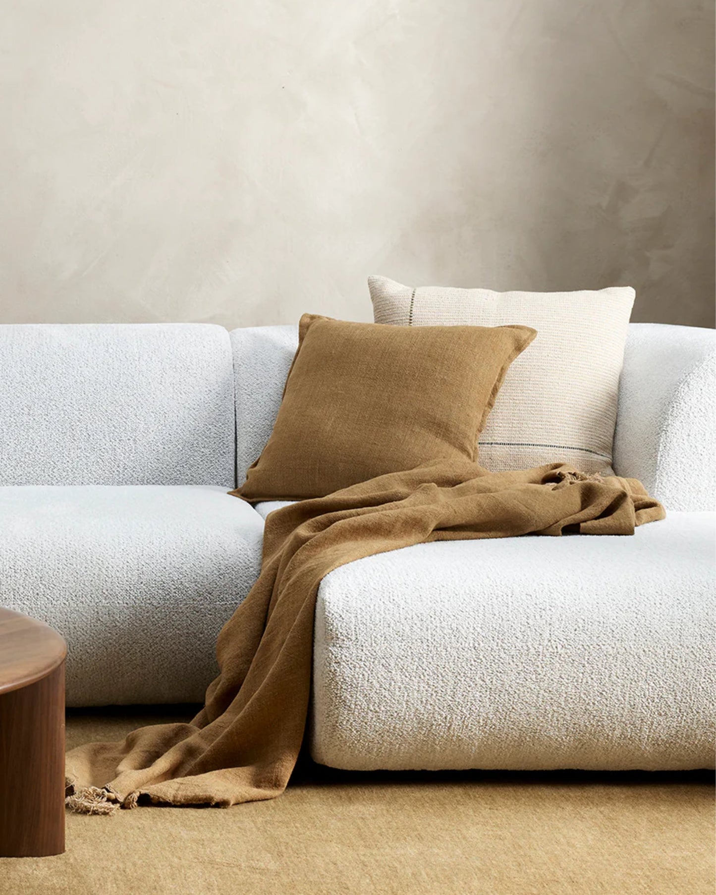 In a contemporary living room scene, a white textured sofa is adorned with FLAXMILL HANDWOVEN LINEN CUSHIONS in beige and white. A light brown throw blanket made from raw linen fibers is elegantly draped over the sofa. To the left, a wooden coffee table partially appears against a wall painted in cloudburst tones.