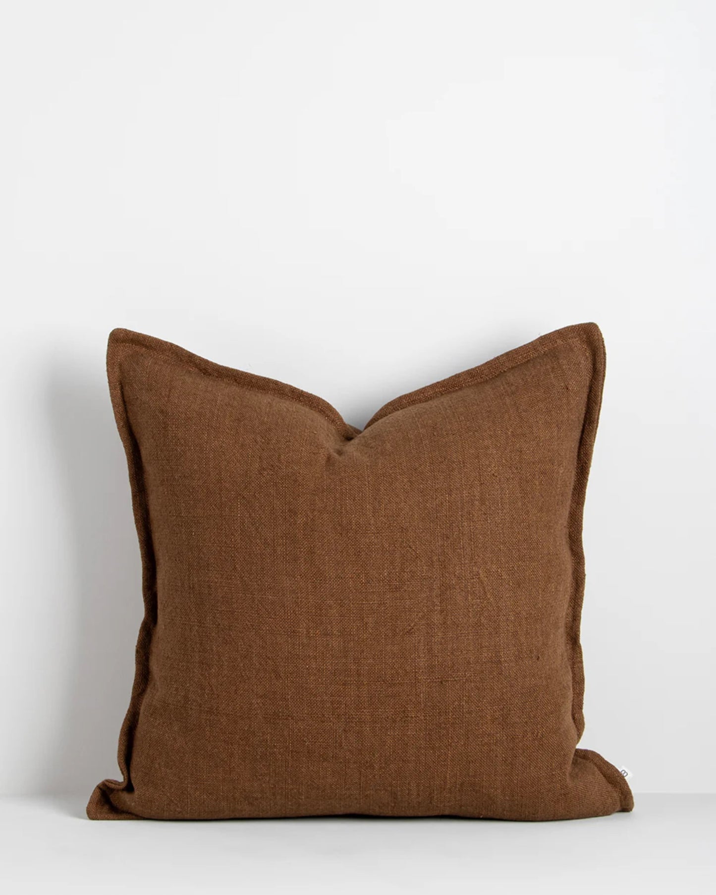 The FLAXMILL HANDWOVEN LINEN CUSHION by Baya, featuring a heavyweight weave and textured brown fabric, is displayed against a plain white background. The cushion's edges are adorned with slightly darker piping, enhancing its defined and structured appearance.