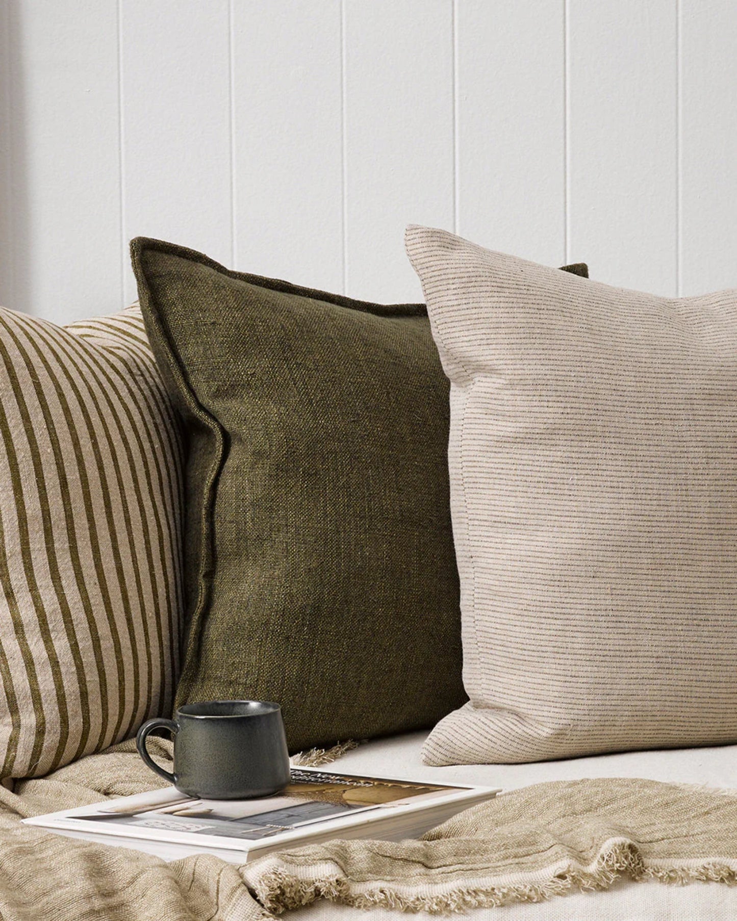 The cozy seating area showcases two FLAXMILL HANDWOVEN LINEN CUSHIONS by Baya, one in a dark green hue and the other in light beige, both crafted from a heavyweight weave. They rest against a wall featuring vertical panels, accompanied by a striped cushion. A folded blanket and a dark mug in cloudburst tone are placed on top of two magazines on the seat.