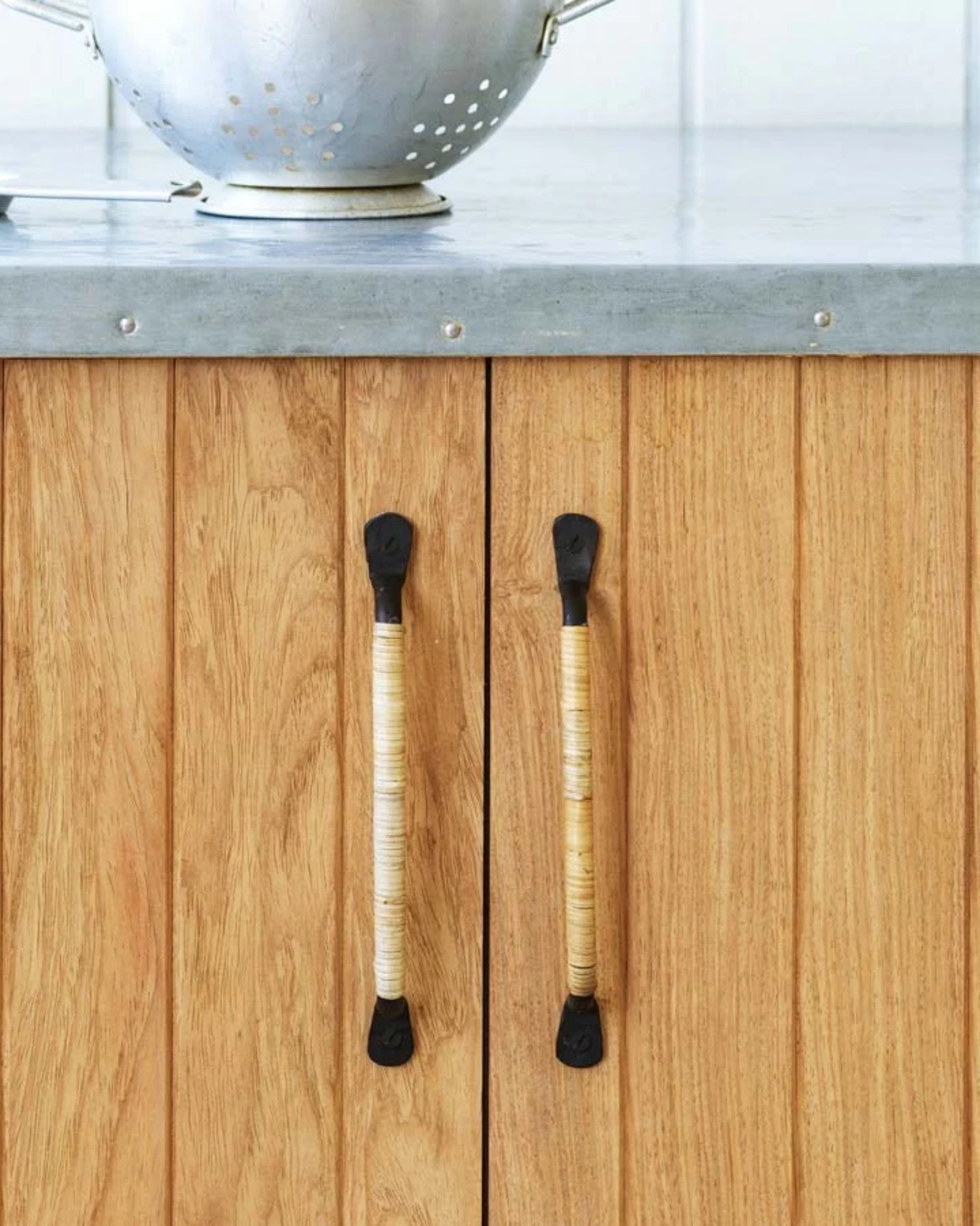 A close-up of a wooden cabinet with vertical panels showcases two FLEET DRAWER PULLs by Society Ink, distinctively wrapped with natural fiber twine. A metal colander sits on the countertop above, which has a smooth, gray finish.