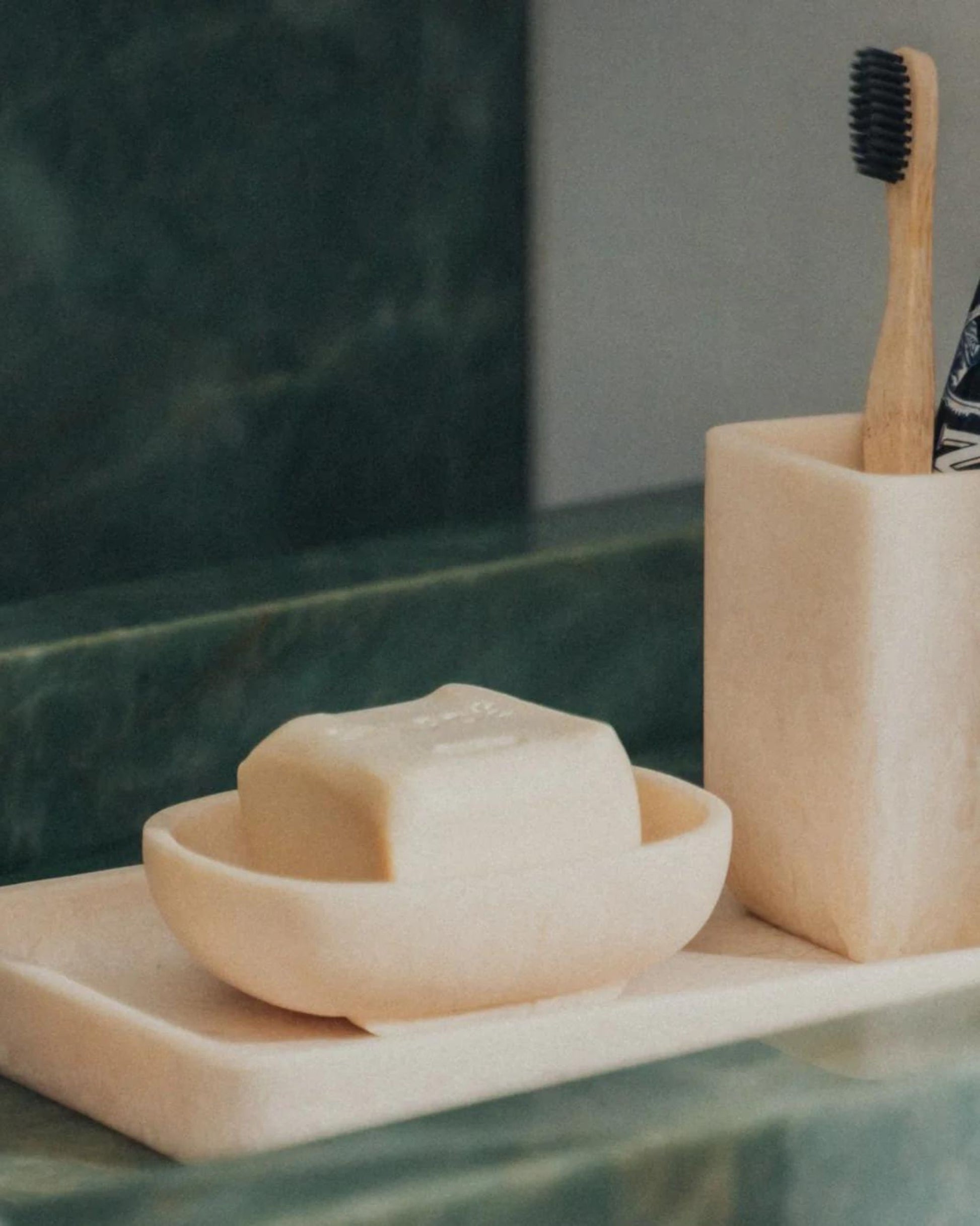 A beige soap bar rests in a Saardé Flow Resin Soap Dish on a sleek tray. Beside it, a toothbrush with a black brush head stands in a matching holder from the Saardé Flow Collection. The dark green marble surface adds an elegant backdrop.