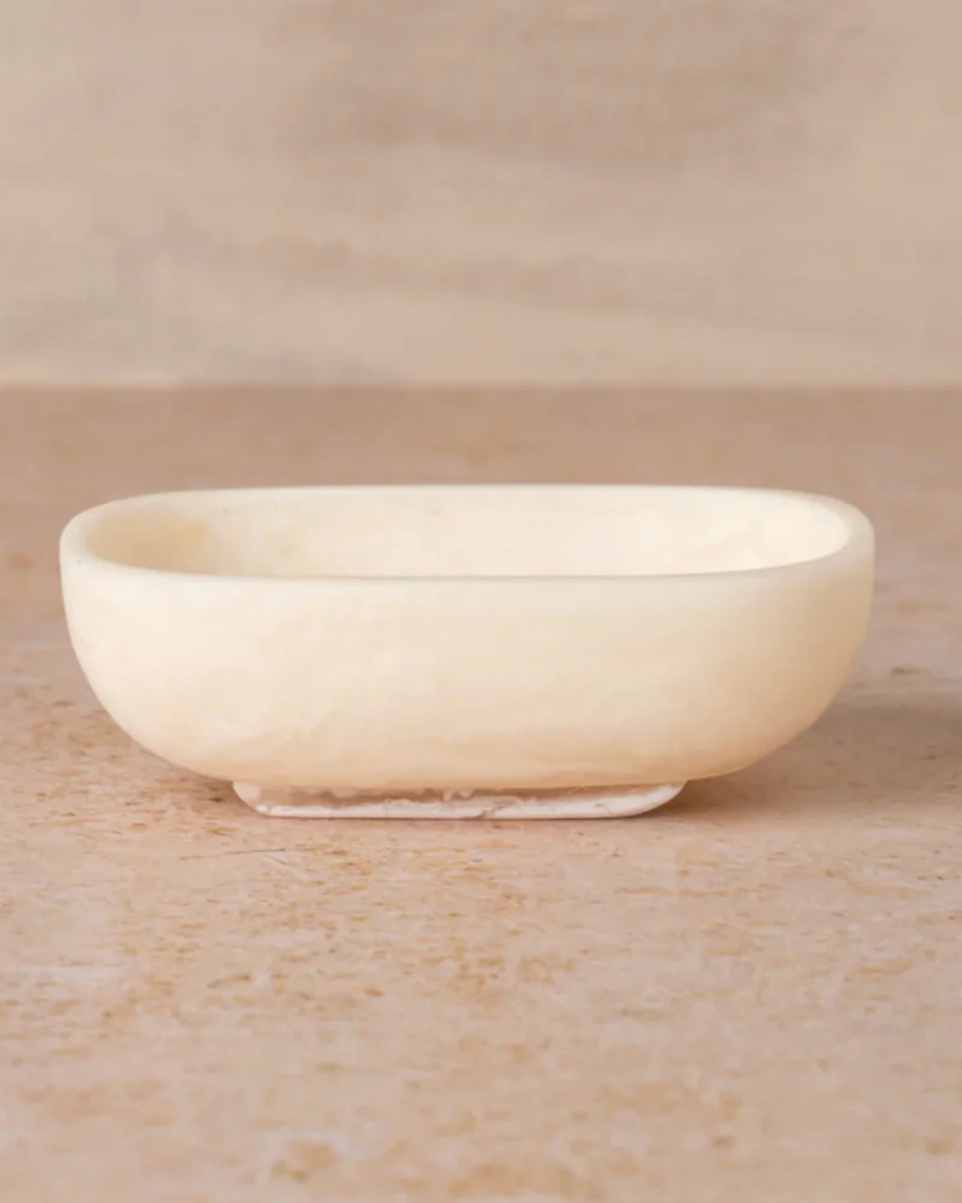 A beige, oval-shaped FLOW RESIN SOAP DISH from Saarde is placed on a marble surface. Featuring smooth edges and a minimalist, elegant design, this handmade soap dish enhances the space with a touch of sophistication.