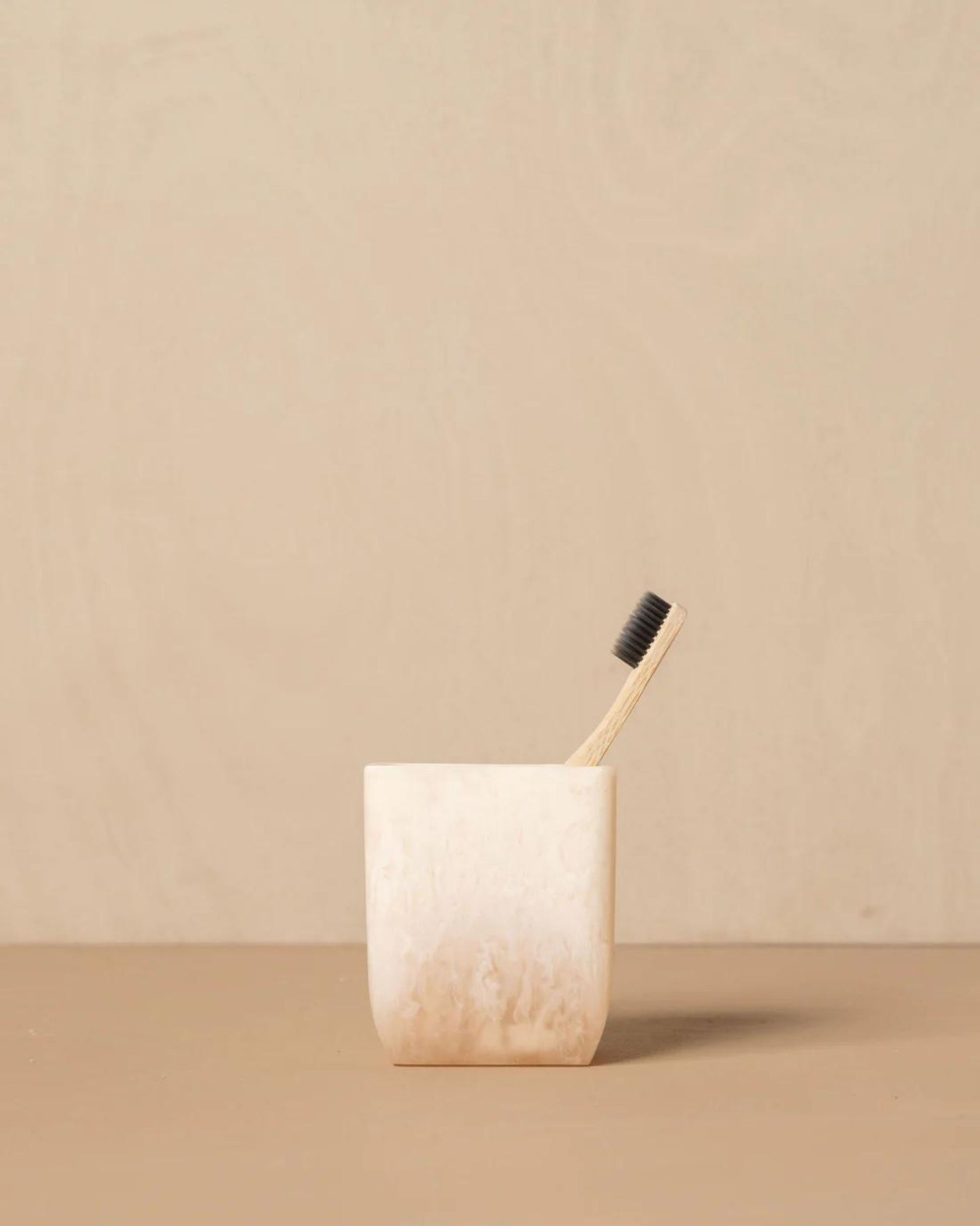 A bamboo toothbrush with dark bristles stands upright in the Saarde FLOW RESIN TOOTHBRUSH HOLDER, an off-white handmade cup showcasing a subtle marble finish, set against a beige background.