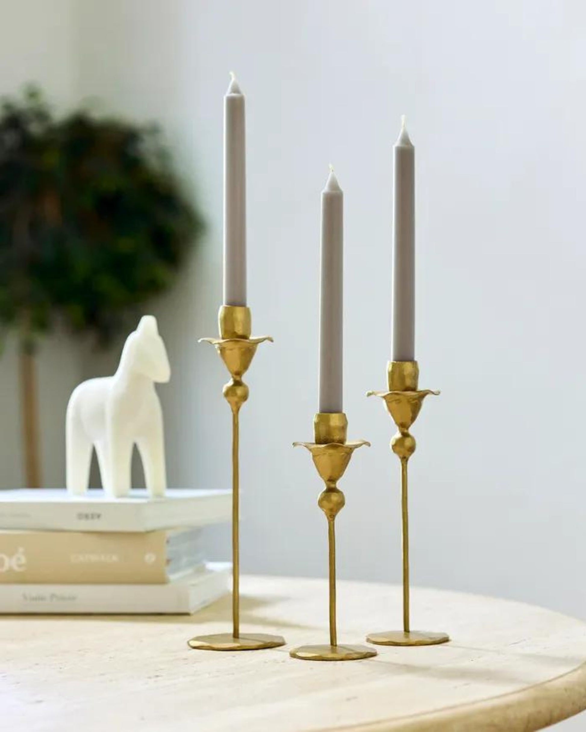 Three sophisticated FLOWER CANDLE HOLDERs by French Country, crafted in gold-forged iron with gray candles, adorn a round table. In the background, two books and a white decorative horse figurine complement the setting, while a potted plant brings a refreshing touch of greenery to this exquisite home décor arrangement.