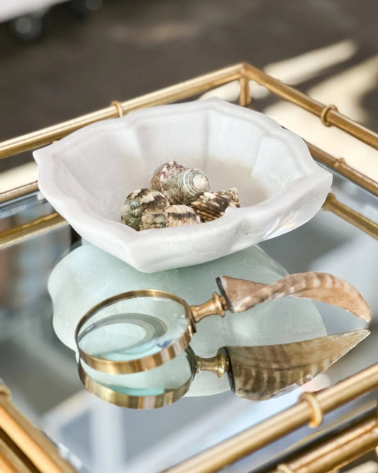 A decorative accent featuring the New direction FLOWER MARBLE BOWLS filled with seashells, paired with a brass-handled magnifying glass and a polished horn, all tastefully arranged on a gold-framed glass table.