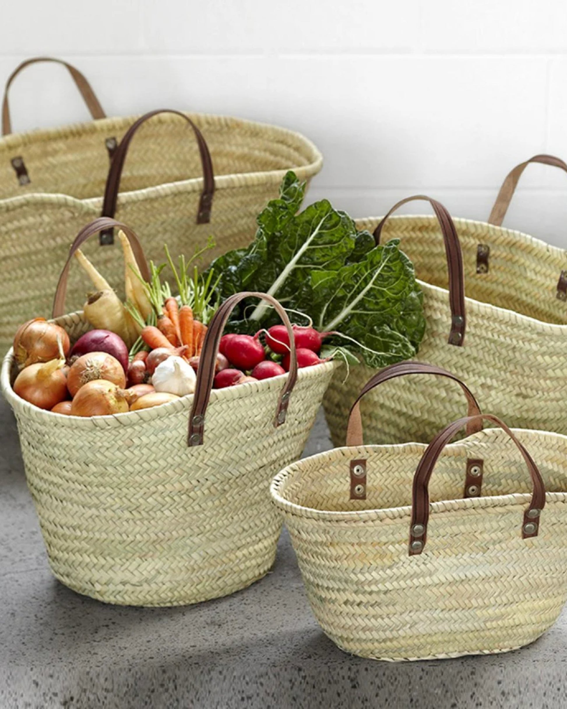 The Epicure Trader's FRENCH MARKET BASKET FLAT HANDLE, with its hand-stitched leather accents, is brimming with fresh produce like onions, carrots, radishes, and leafy greens and elegantly presented on a gray surface.