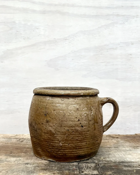 A FRENCH VINTAGE EARTHENWARE PRESERVING POT by All Things French, featuring a rustic brown ceramic design with a handle, is placed on a textured wooden surface. Its weathered appearance exudes the vintage charm of South West France, reminiscent of traditional rustic earthenware.
