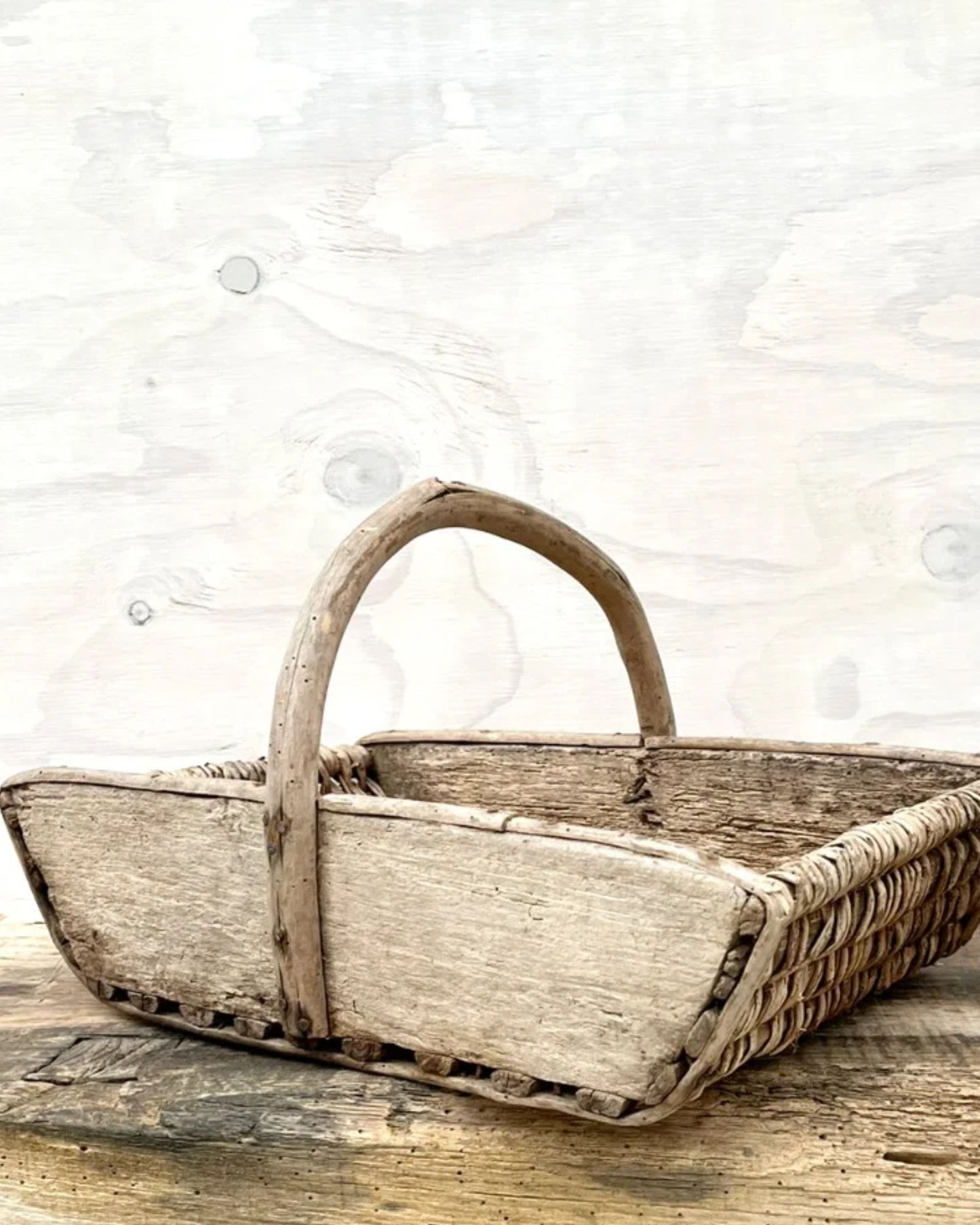 A charming French Vintage Wooden Trug by All Things French, featuring a tall, curved handle, rests on a wooden surface. Its woven base and weathered texture add to its antique appeal, making it an ideal piece for rustic décor. In the background is a light wooden wall.