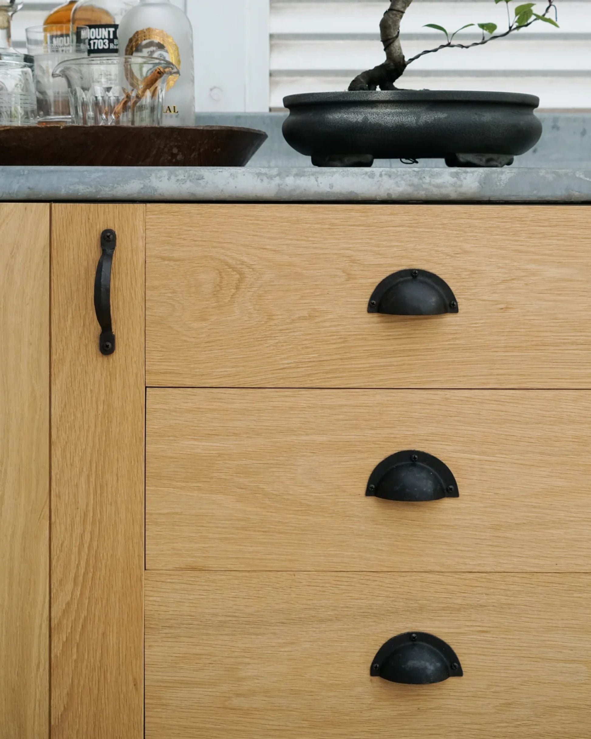 This wooden cabinet, crafted with the GALLEY DRAWER PULL by Society Ink, features three drawers adorned with sleek black pulls. The top surface is elegantly arranged with glasses and a bottle of Mount Gay rum, complemented by a small bonsai tree in a black pot—ideal for capturing the essence of a ship's galley kitchen.