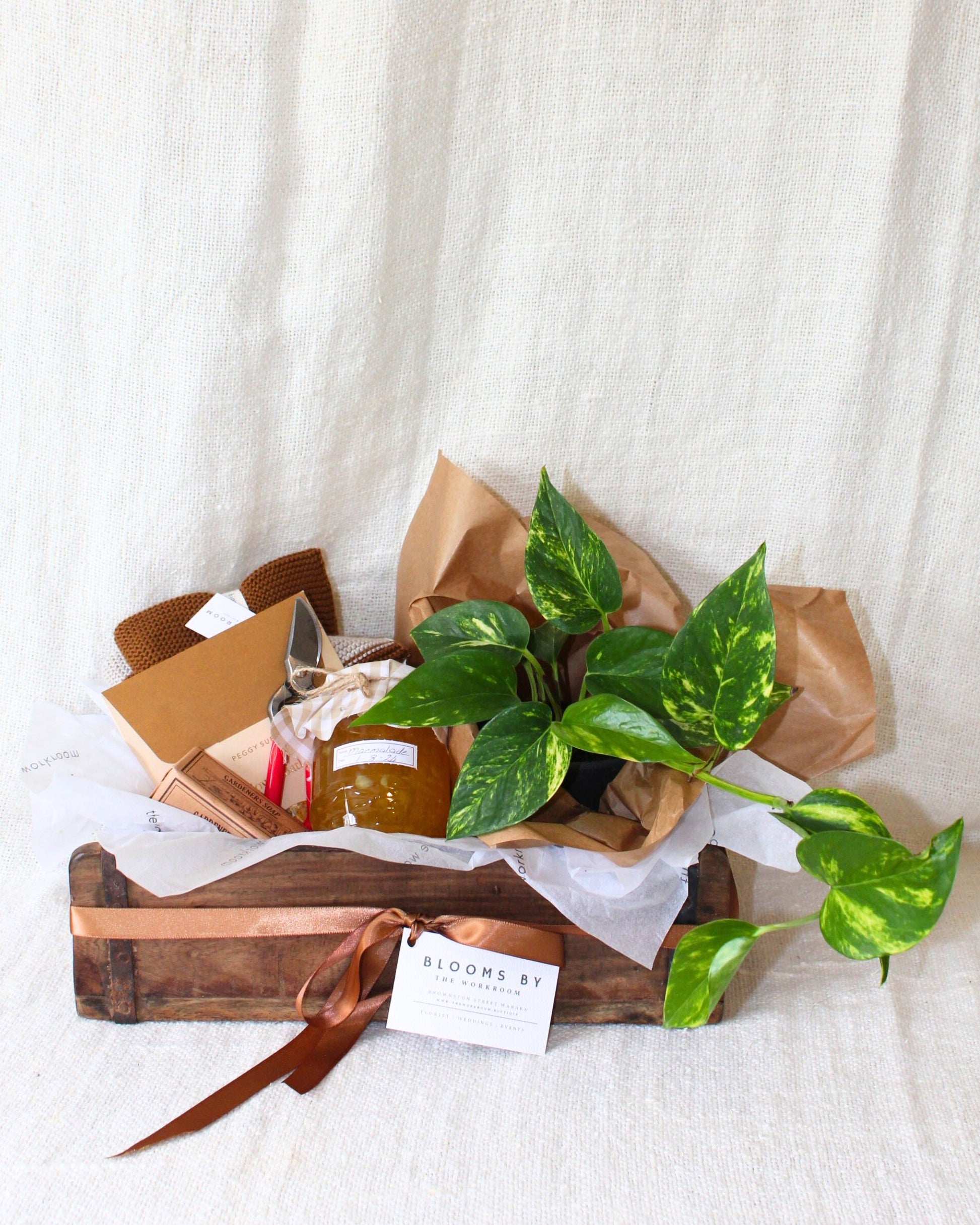 The GARDEN LOVERS' GIFT BASKET by The Workroom NZ features a rustic wooden crate inspired by a vintage brick mould. Inside, you'll find a potted green plant, a jar of jam, a honey dipper, cozy warm brown socks, scissors, and an elegantly ribbon-adorned business card, all beautifully set against a beige fabric backdrop. It's the perfect gift for those who appreciate nature's charm.