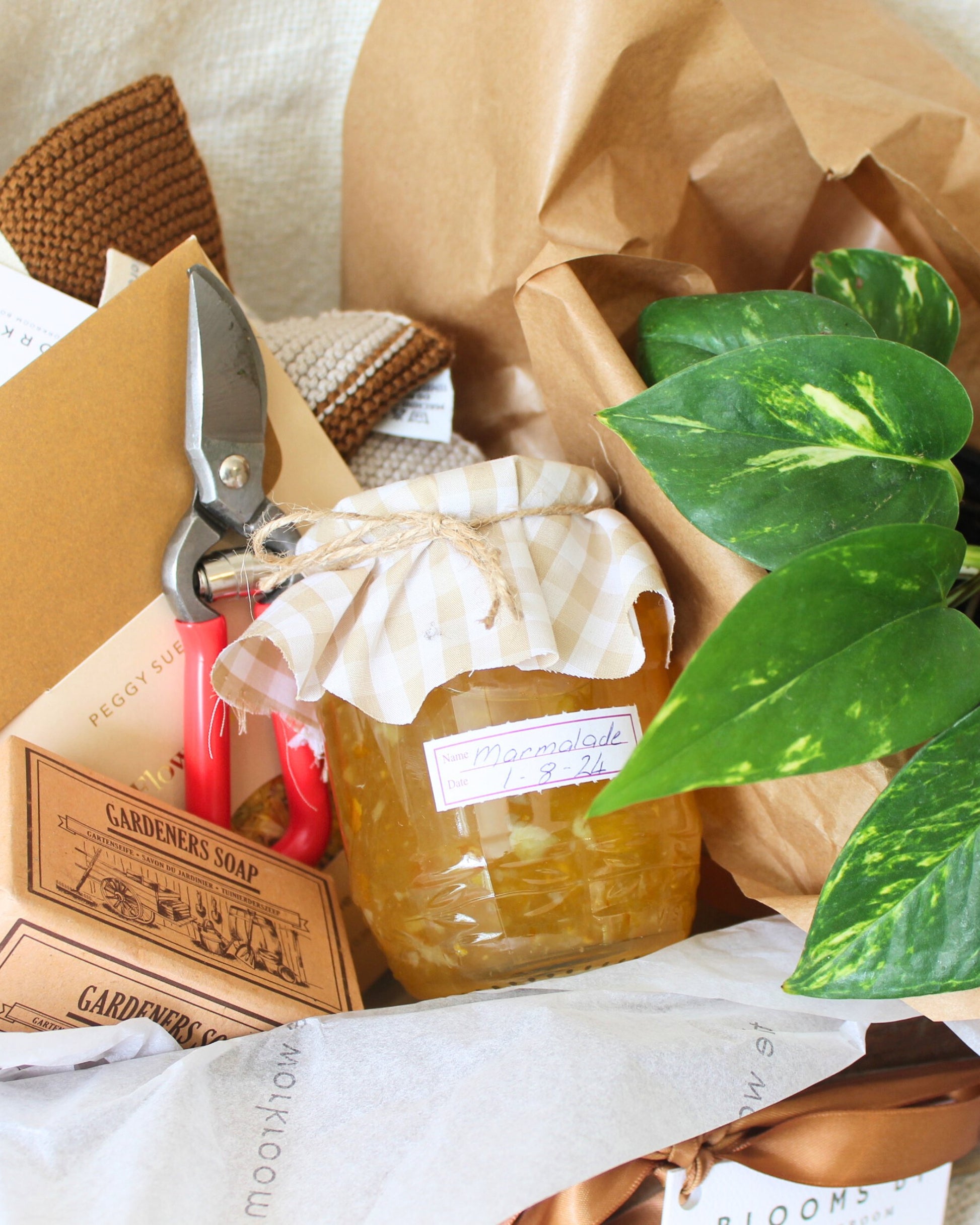 Introducing the GARDEN LOVERS' GIFT BASKET by The Workroom NZ: a charming collection including a jar of marmalade with a checkered lid, essential gardening tools like garden pruners, a bar of gardener's soap, a lush green-leafed plant, and an engaging book. This delightful ensemble is elegantly displayed on white and nestled in brown paper for that perfect rustic touch.