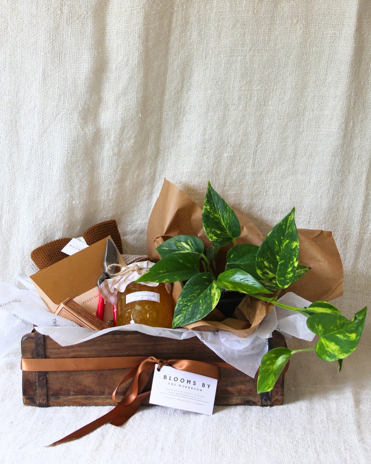 The GARDEN LOVERS' GIFT BASKET from The Workroom NZ features a charming wooden box containing a lush green leafy plant, a jar of honey, and a beeswax candle. Accompanied by matches and brown paper-wrapped gardening tools, it is elegantly adorned with a ribbon and card displaying the name Blooms by. This ensemble is set beautifully against a beige textured fabric backdrop.