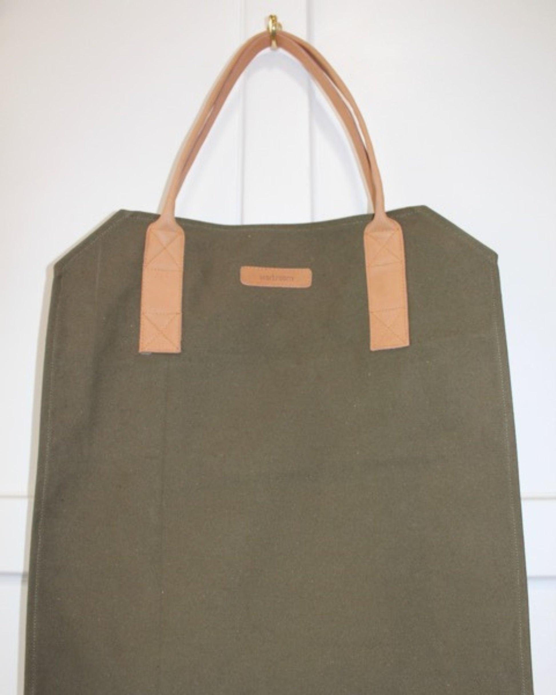 The GARDENER’S CANVAS CARRY ALL by The Workroom, a large olive green tote bag with tan leather handles, hangs on a white wall. This versatile carry-all is designed in a simple rectangular shape with two sturdy handles, making it perfect for carrying essentials.