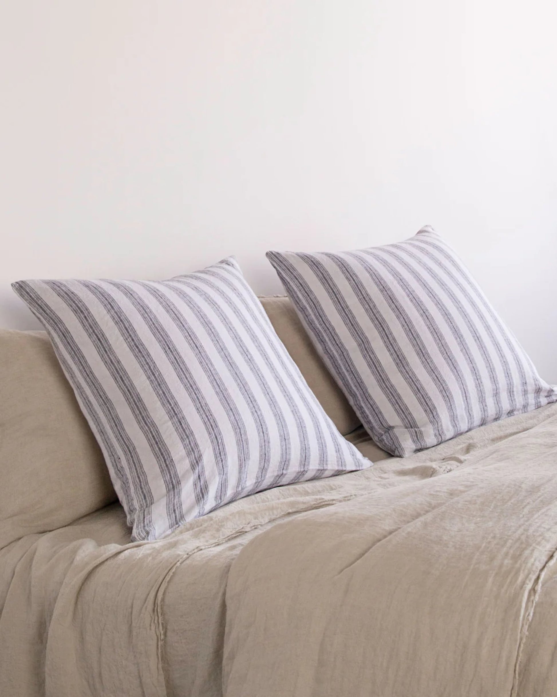 Two Basix Stripe Euro pillowcases from Hale Mercantile Co are placed on a light beige bed, adorned with a soft, textured blanket. The backdrop features a plain white wall that enhances the minimalist and cozy atmosphere.