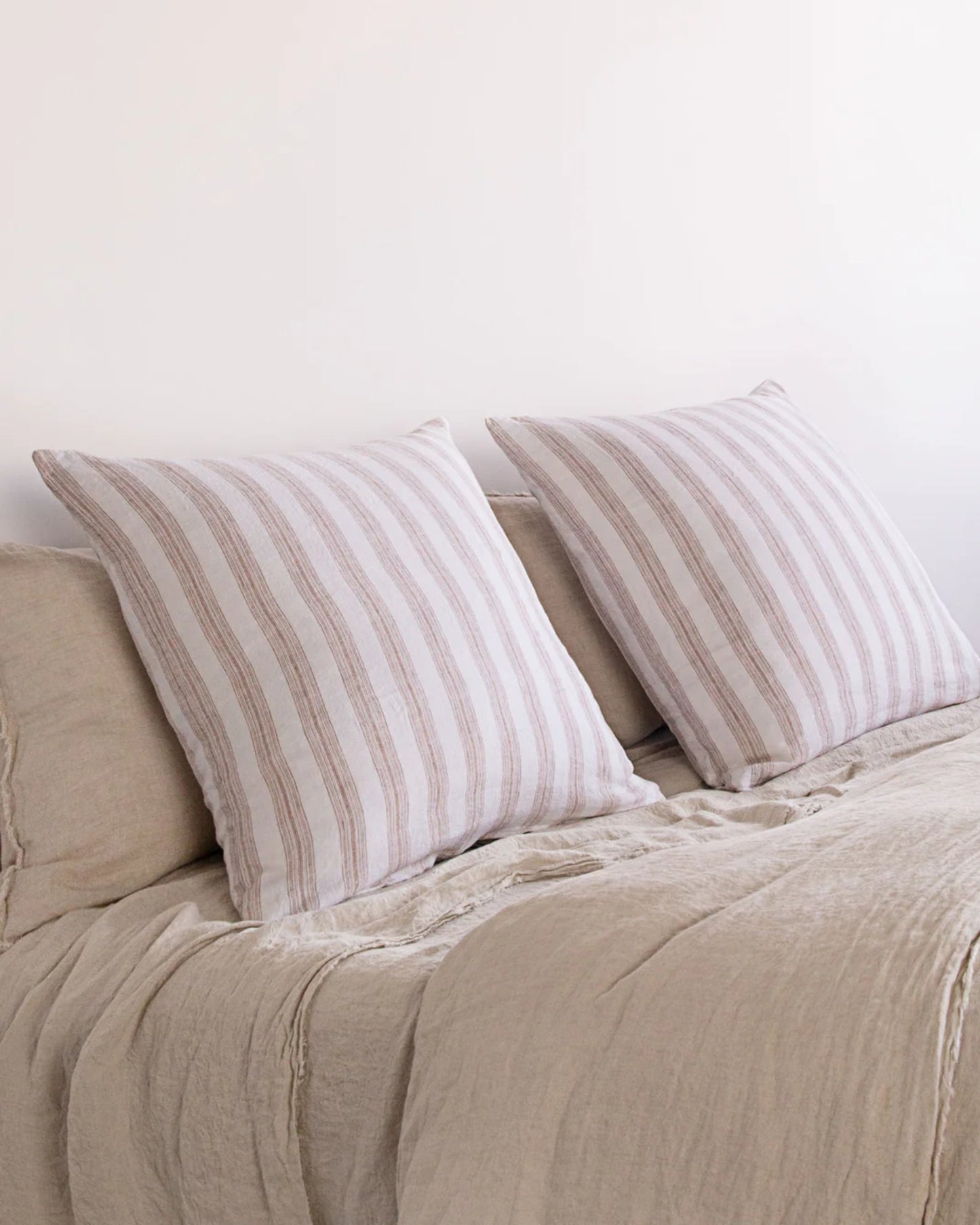 Two beige pillows made from Hale Mercantile Co's HALE BASIX STRIPE EURO PILLOWCASE, featuring brown stripes, rest on a beige linen-covered sofa. These European pillowcases enhance the minimalistic setting, creating a cozy and neutral atmosphere.