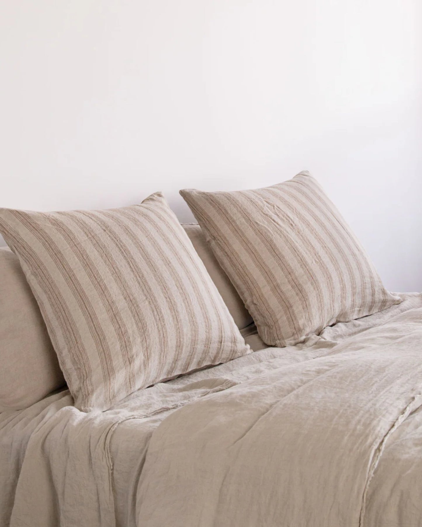 A neatly made bed features light beige, textured pure linen bedding. Two large, square HALE BASIX STRIPE EURO PILLOWCASES by Hale Mercantile Co rest against a plain white wall, creating a calm and minimalist aesthetic.