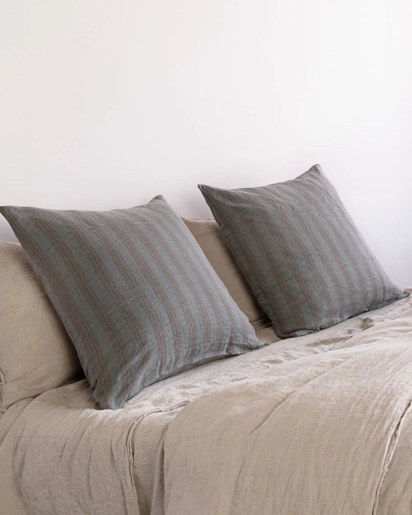 Two large throw pillows in gray, featuring Hale Mercantile Co's HALE BASIX STRIPE EURO PILLOWCASE, rest against the back of a beige sofa or daybed. The pure linen texture of the fabric enhances a cozy and minimalist atmosphere against a plain white wall.