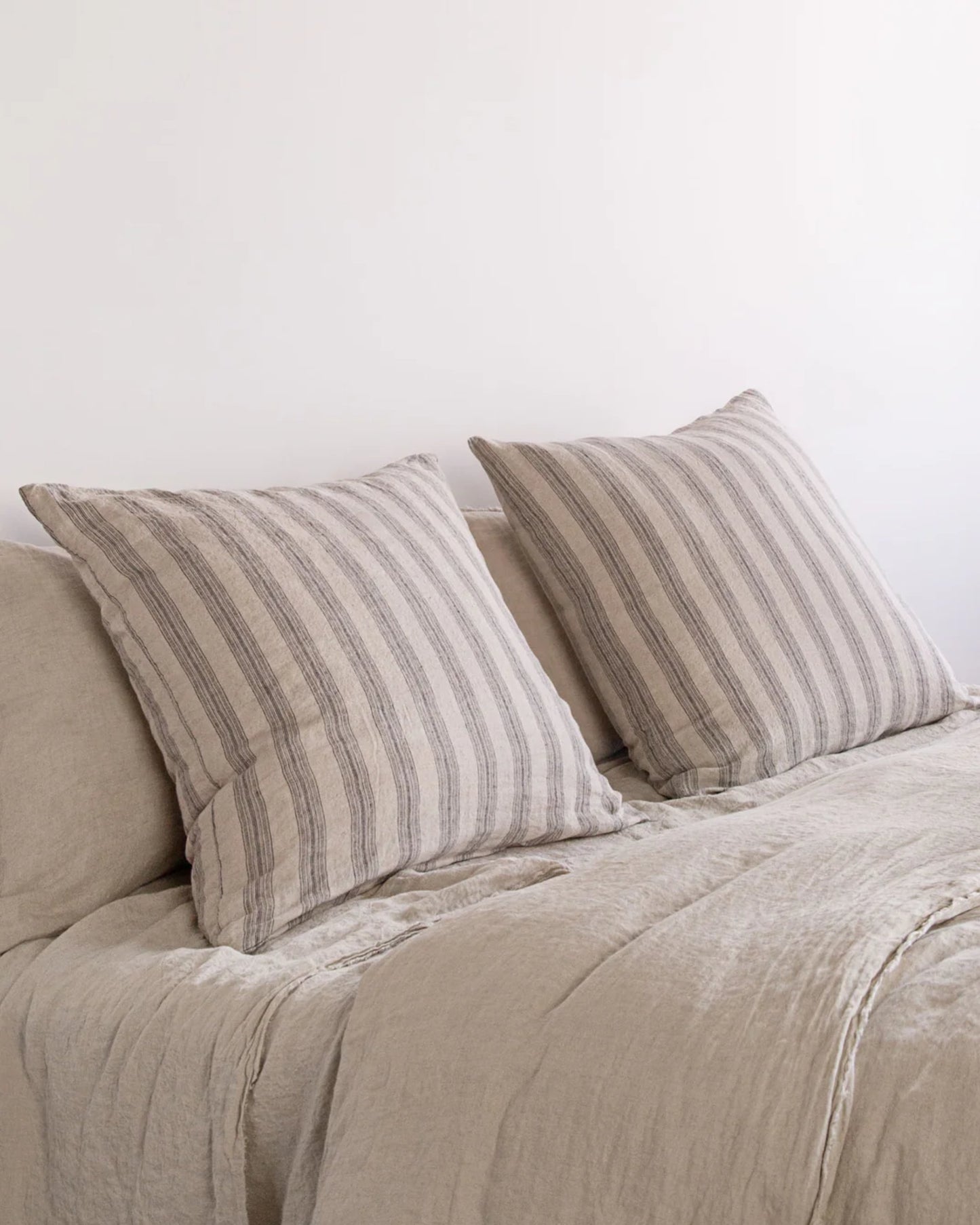 A cozy bed with beige, striped pillows and matching Hale Mercantile Co's Basix Stripe Linen bedding against a plain white wall. The HALE BASIX STRIPE EURO PILLOWCASE fabric appears soft and textured, creating a relaxed and inviting atmosphere.