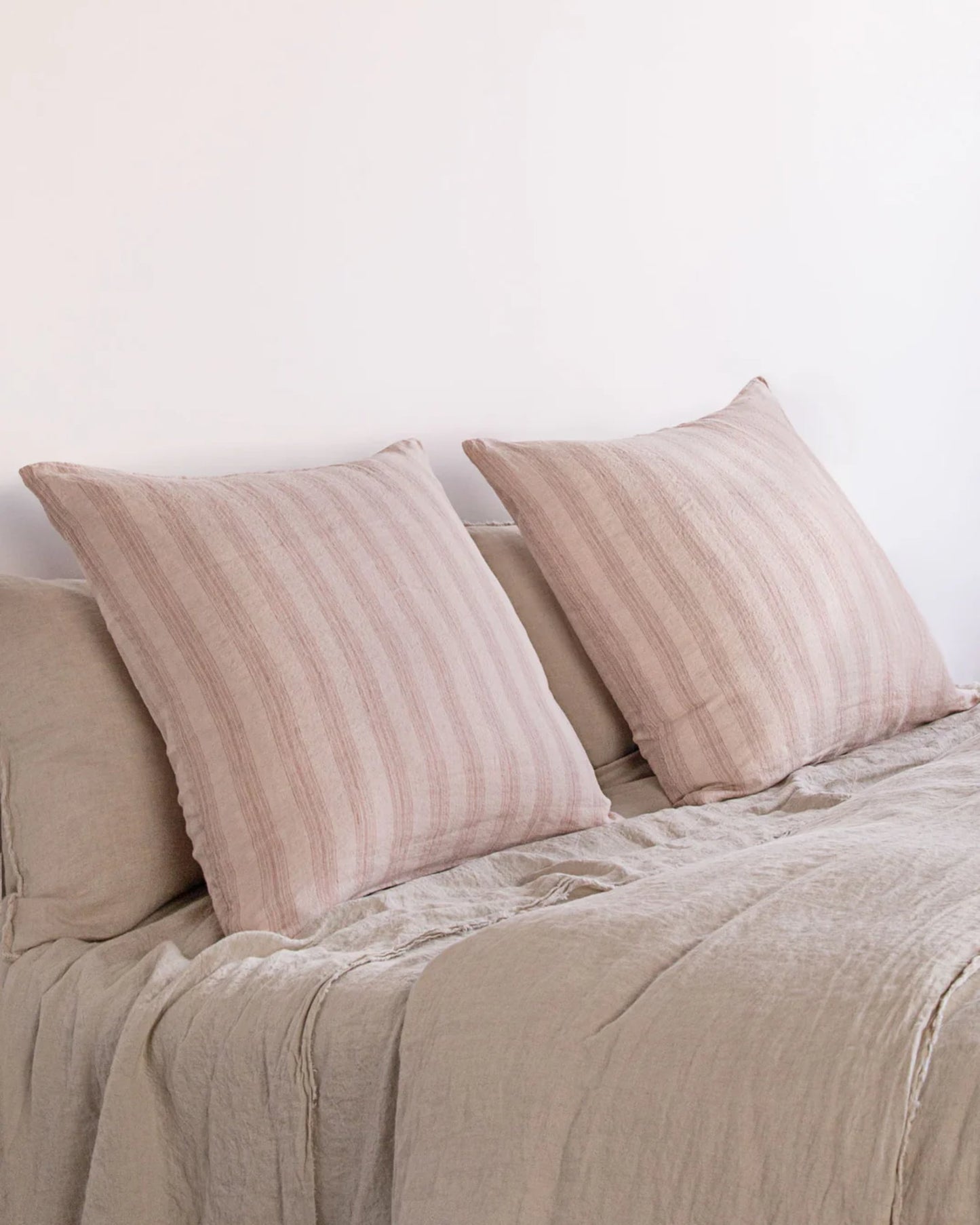 Two HALE BASIX STRIPE EURO PILLOWCASEs in beige, designed with subtle stripes, rest against a plain white wall on a bed covered with a matching textured bedspread. This minimalistic and calming setting is gracefully accented by pure linen elements from Hale Mercantile Co.