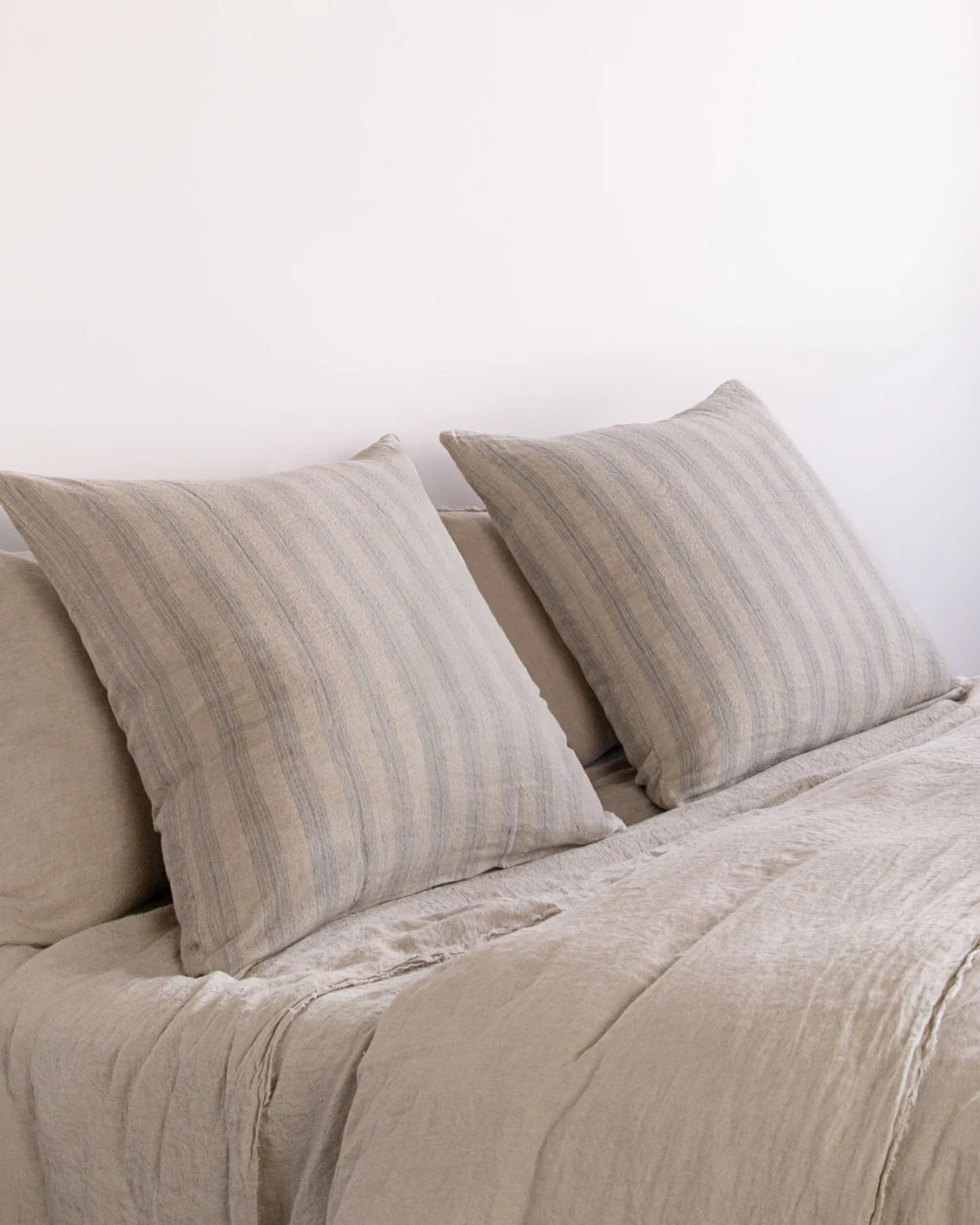 A minimalist bedroom features a neatly made bed adorned with Basix Stripe pillows and a matching bedspread, all set against a plain white wall. The pure linen bedding exudes softness and allure, creating a serene, neutral-toned space accentuated by elegant Hale Mercantile Co.'s HALE BASIX STRIPE EURO PILLOWCASES.