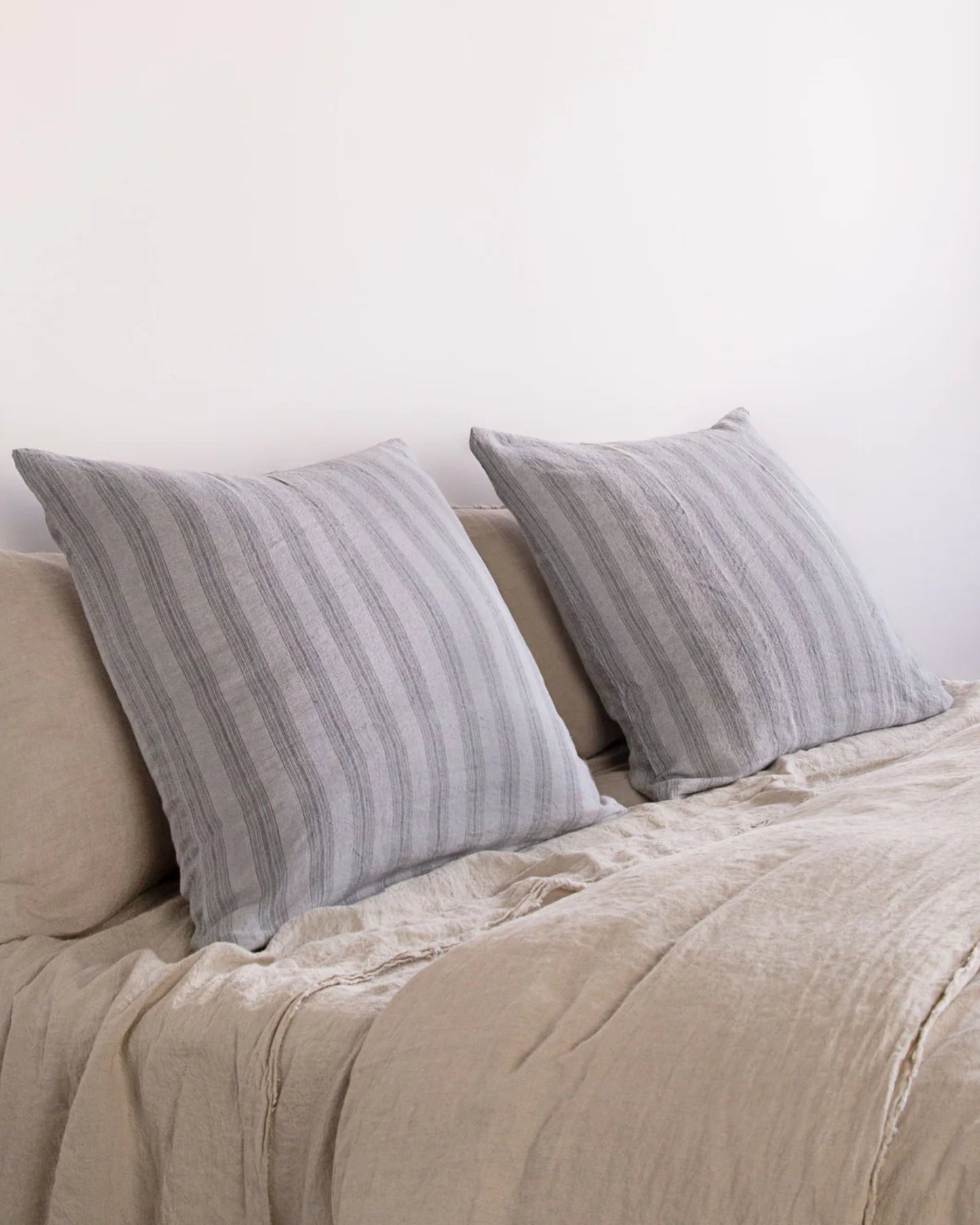 Two large pillows covered with HALE BASIX STRIPE EURO PILLOWCASE by Hale Mercantile Co rest on a beige bedspread. The bed is set against a plain white wall, highlighting the minimalist charm and creating a cozy bedroom setting.