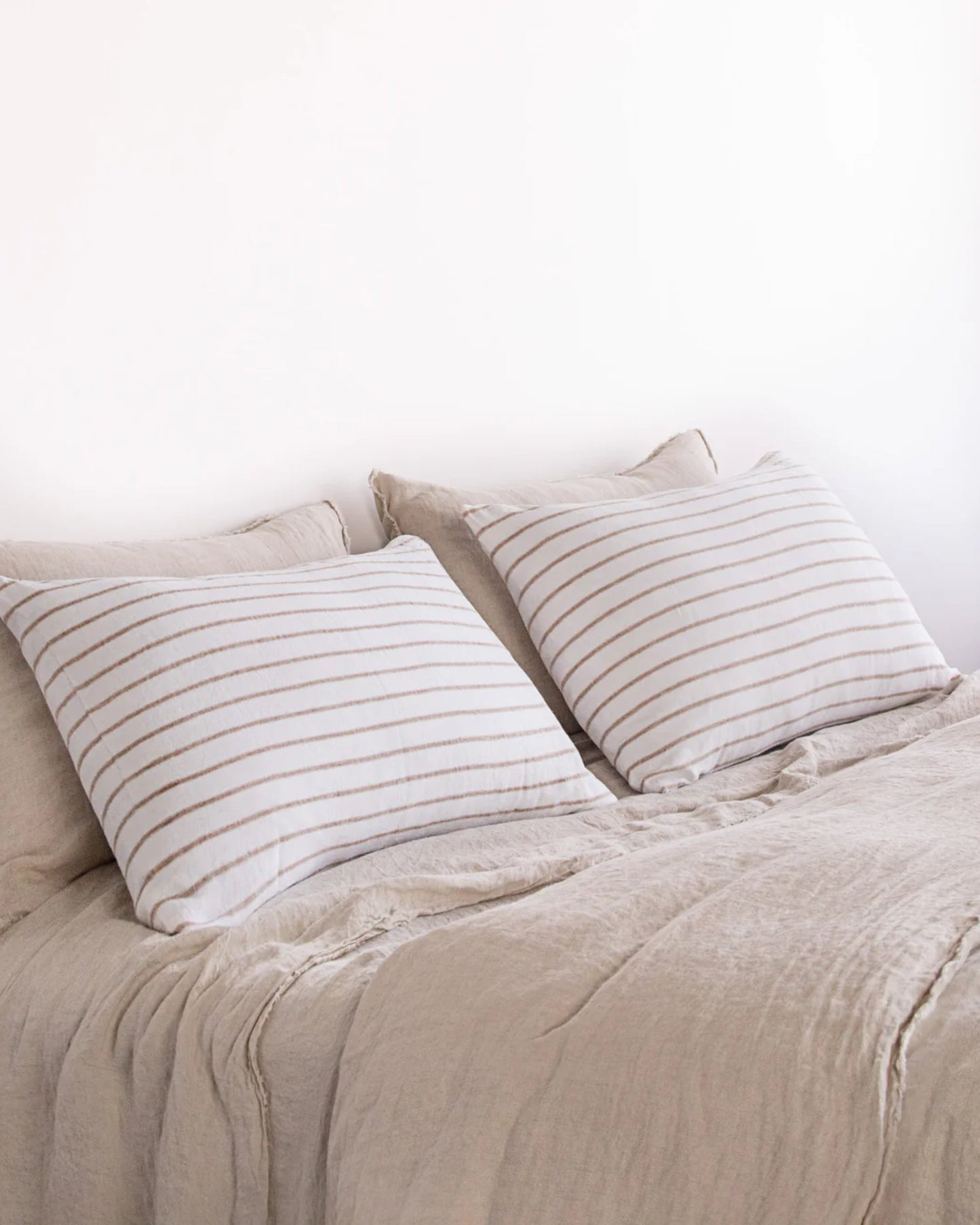 A neatly made bed features two large pillows adorned with thin, horizontal beige stripes. The HALE BASIX STRIPE PILLOWCASE by Hale Mercantile Co brings a touch of elegance, while light brown pure linen sheets and a duvet complete the minimalist and cozy setting against a plain white wall backdrop.