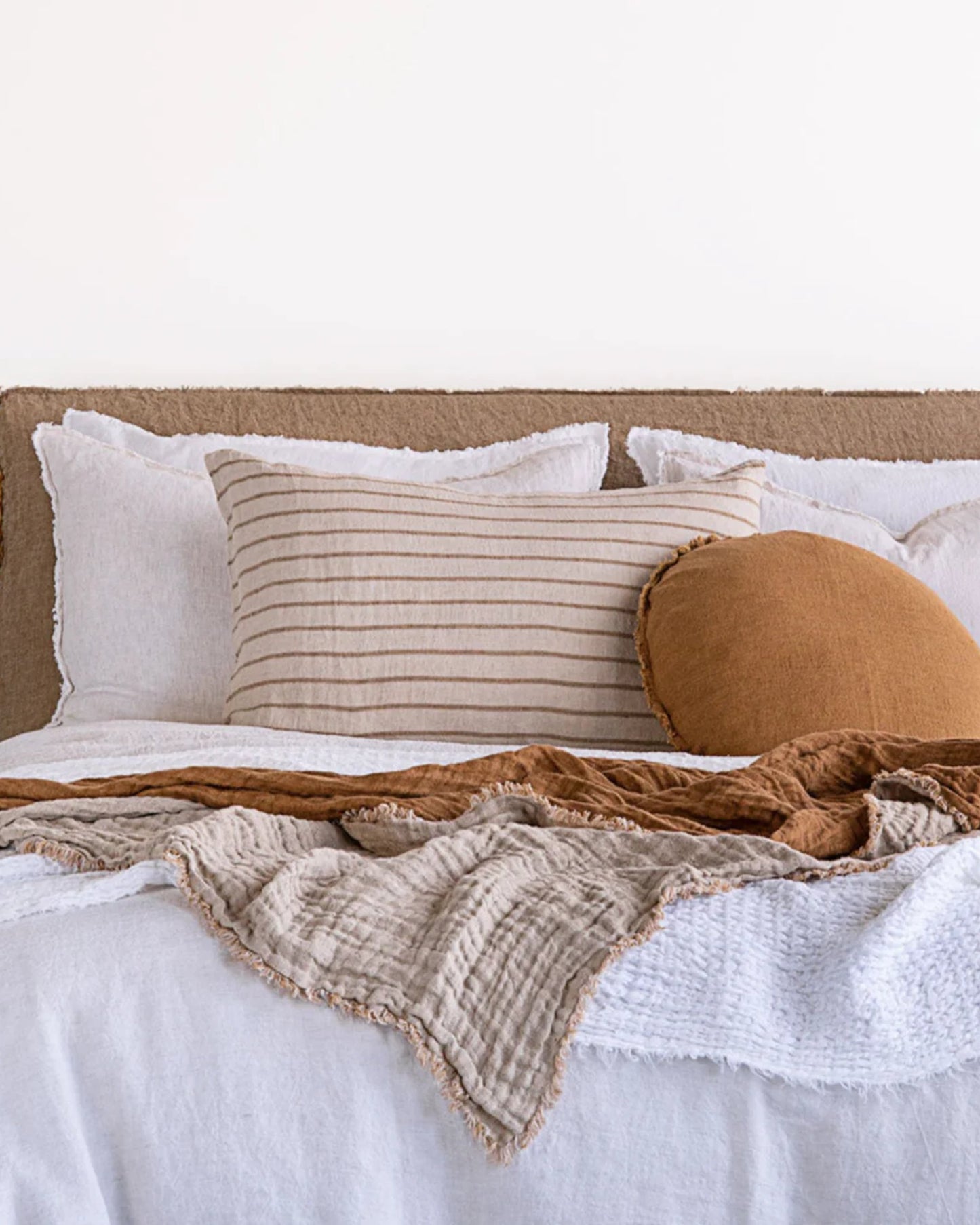 A cozy bed with layered white and beige linen bedding, featuring a Hale Mercantile Co HALE BASIX STRIPE PILLOWCASE and a round brown pillow. A textured beige blanket is draped over the white comforter, creating a warm and inviting atmosphere.