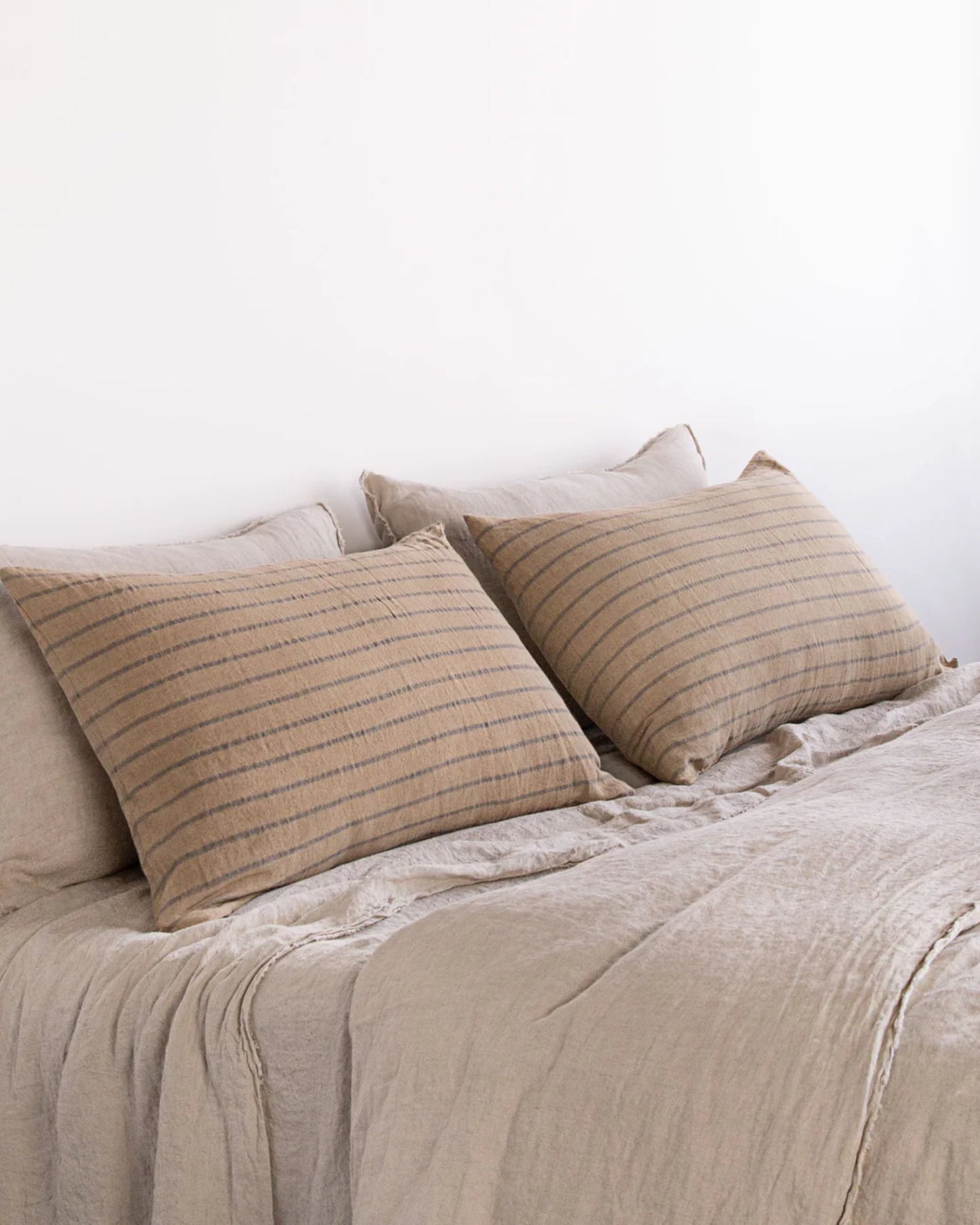 A neatly made bed features pure linen bedding in light tan, complemented by two large Hale Mercantile Co HALE BASIX STRIPE PILLOWCASES. The subtle striped patterns contribute to a minimalist, serene atmosphere against the white wall backdrop.