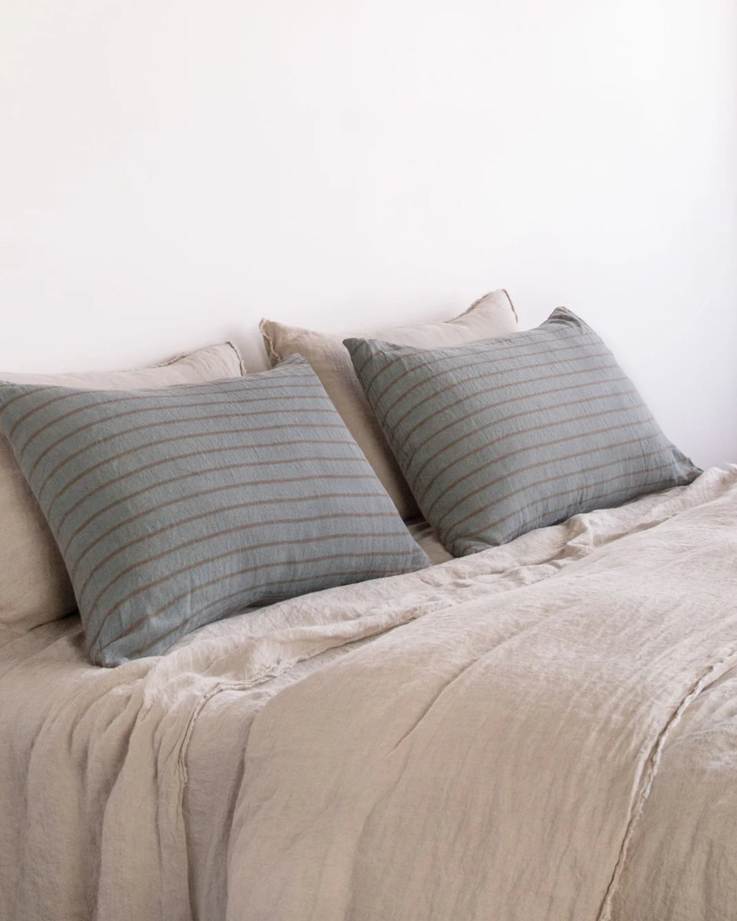 A neatly made bed featuring two Hale Mercantile Co Basix Stripe Pillowcases in muted tones. The bedspread is crafted from pure linen, showcasing a soft, textured fabric in a neutral color that perfectly complements the simple and serene bedroom decor.