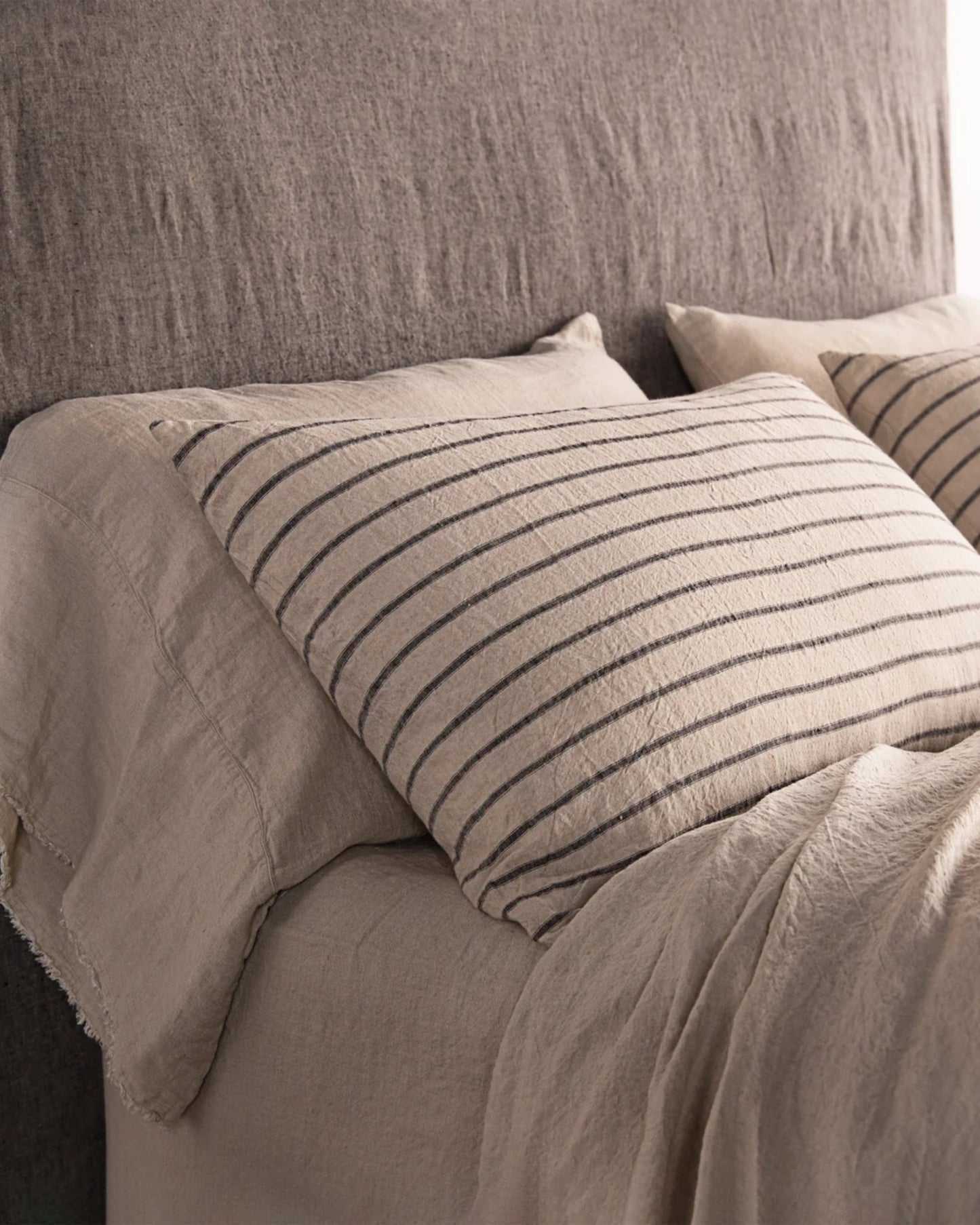 A cozy bed is neatly arranged with beige sheets and features a brown linen headboard. Two pillows sit side by side, one in a HALE BASIX STRIPE PILLOWCASE by Hale Mercantile Co and the other matching the sheets. The pure linen bedding showcases a soft, textured appearance.