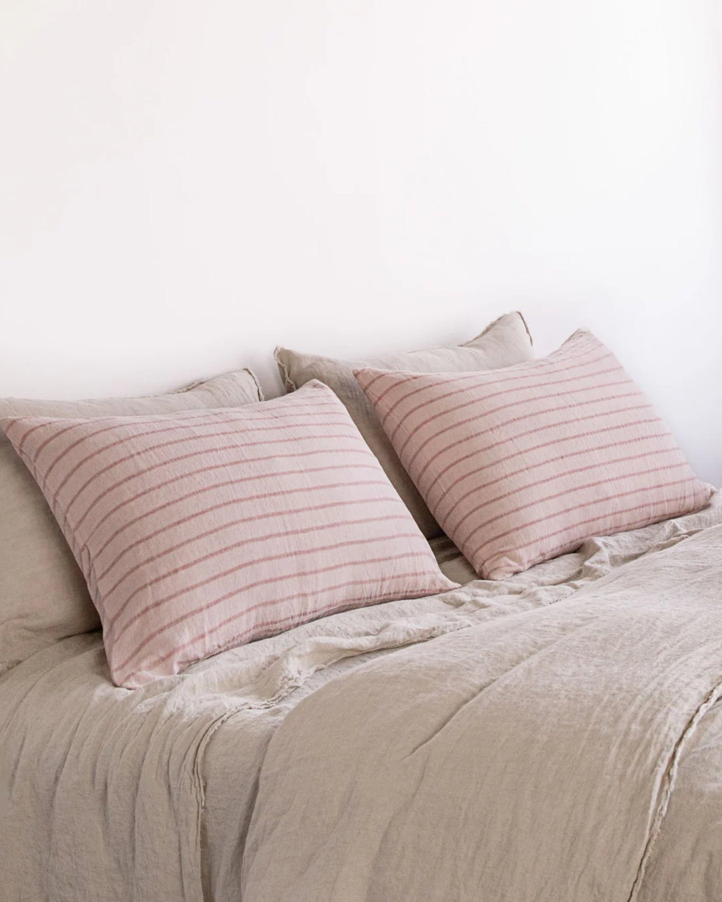 A neatly made bed with beige pure linen sheets and two large Hale Mercantile Co HALE BASIX STRIPE PILLOWCASES. The pillowcases feature a subtle pink and beige striped pattern, perfectly complementing the neutral color scheme of the bedding and room.