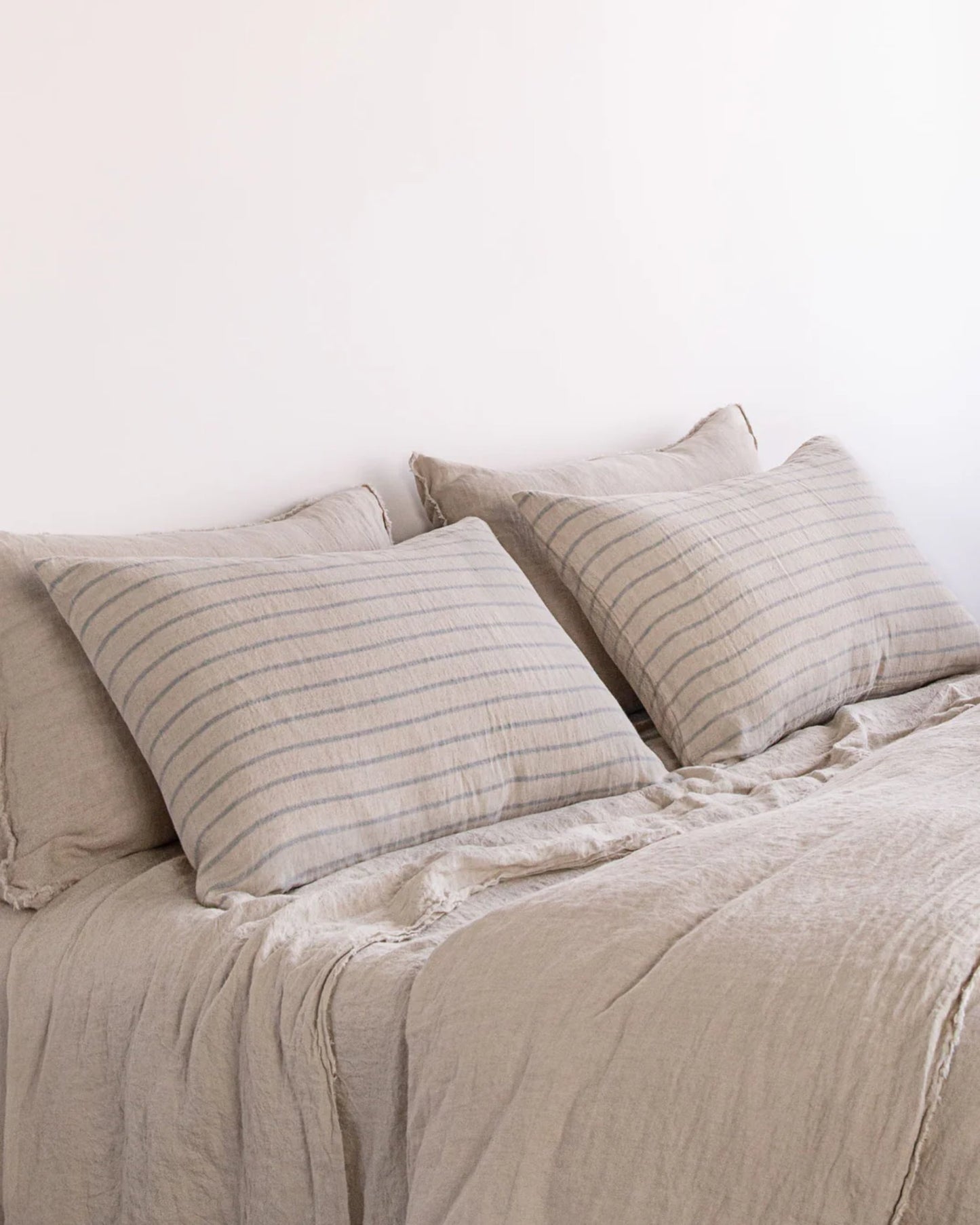 A neatly made bed features beige, pure linen bedding with two large Hale Basix Stripe Pillowcases from Hale Mercantile Co, characterized by subtle horizontal stripes. The soft, textured appearance enhances the minimalistic setting, creating an inviting contrast against the plain white wall.