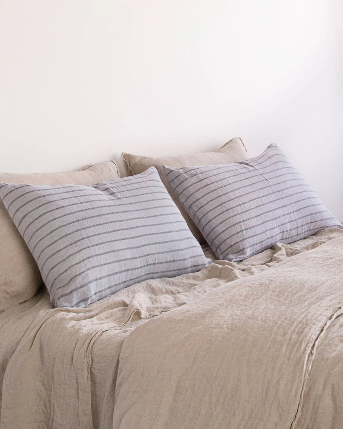 The bed is beautifully arranged with pure linen bedding and two large Hale Mercantile Co's HALE BASIX STRIPE PILLOWCASES boasting gray and white stripes, standing out against a plain white wall. These soft linens contribute to an inviting, serene, and cozy atmosphere.