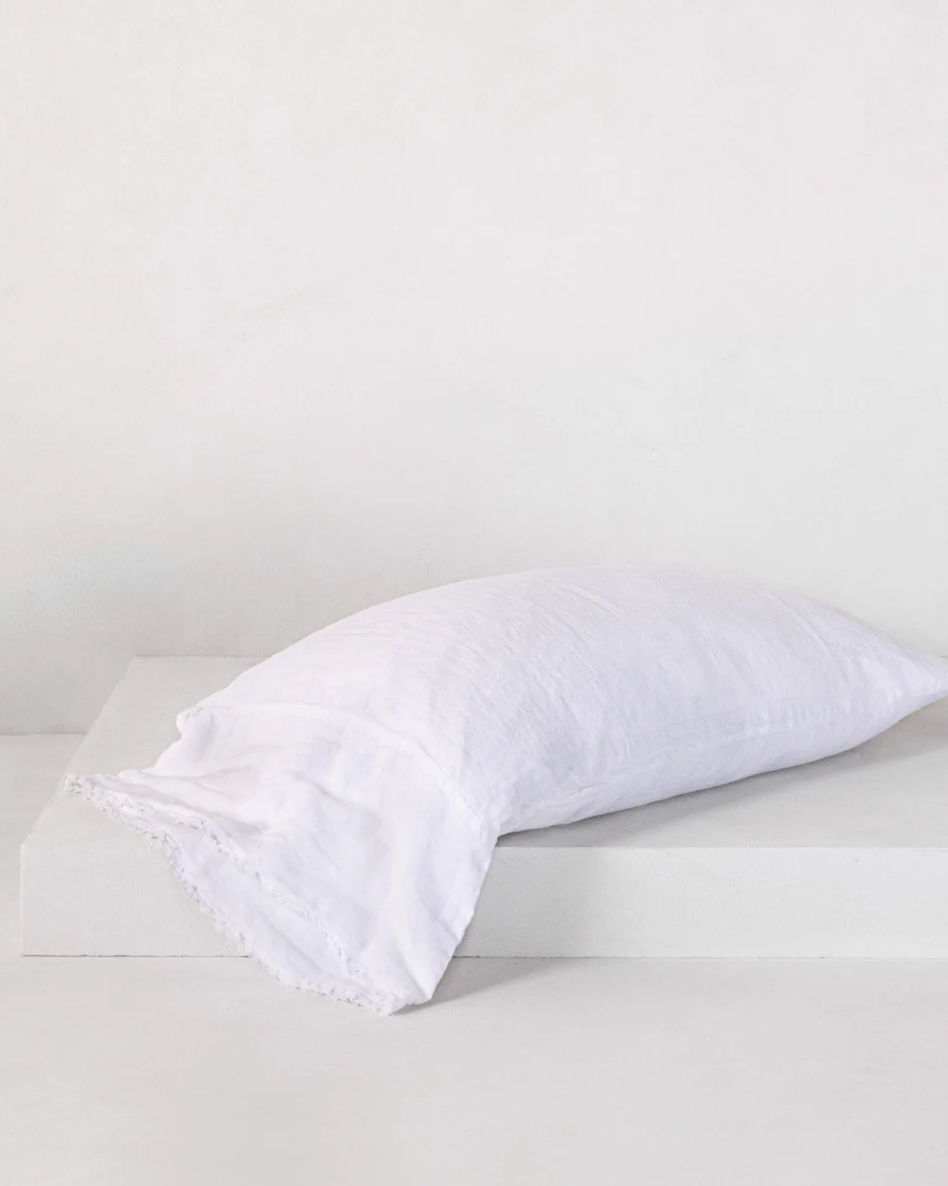 A crisp white pillow with the HALE FLOCCA FLOPPY END PILLOWCASE by Hale Mercantile Co rests on a light, textured surface featuring subtle fringed edges, creating a serene and minimalist aesthetic. The background is a simple off-white wall, enhancing the Flocca hand-tufted edges for a clean and fresh appearance.