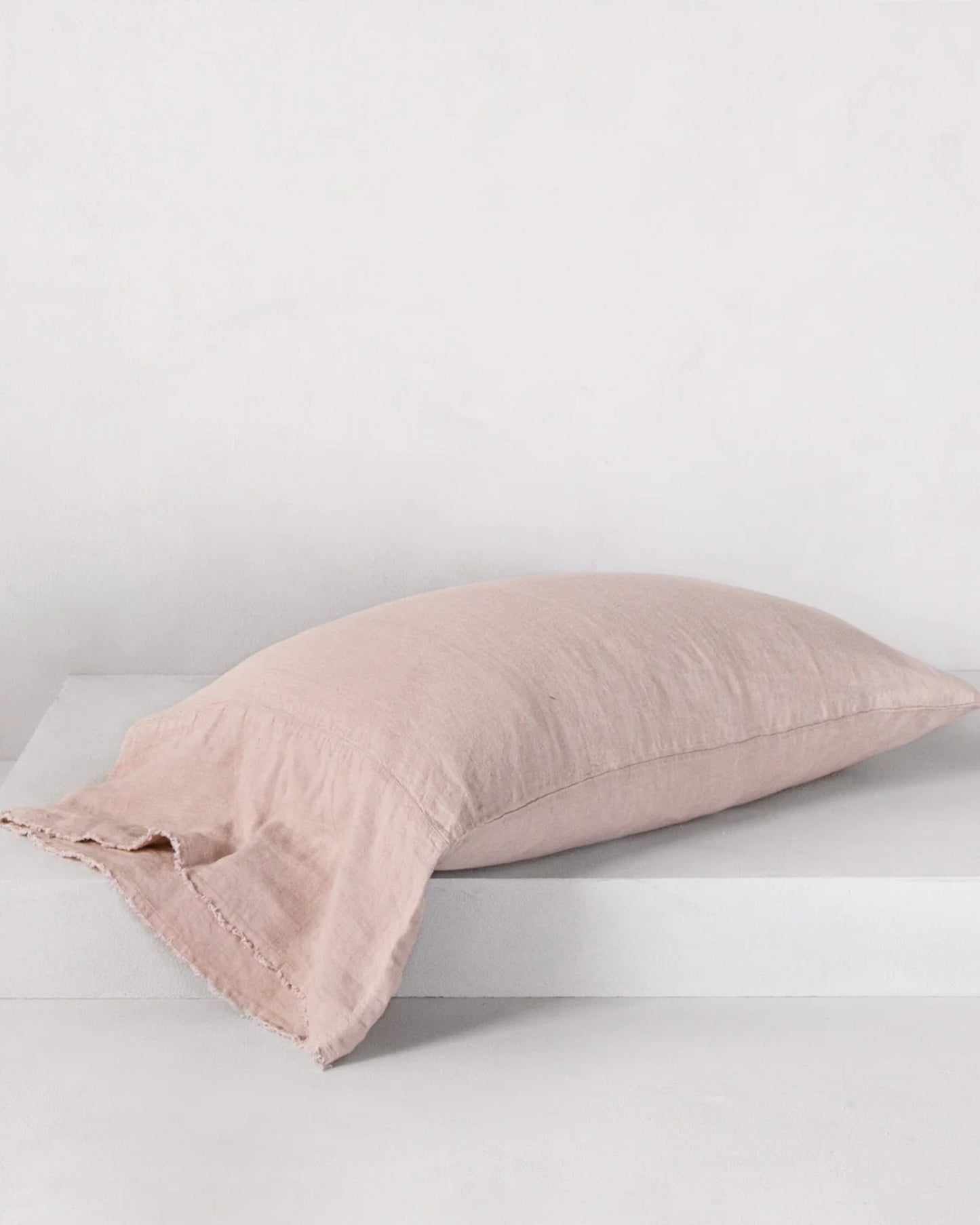 The HALE FLOCCA FLOPPY END PILLOWCASE by Hale Mercantile Co, featuring a soft pink hue and linen texture with Flocca hand-tufted edges, rests on a white surface against a light gray background. Its fabric has a slightly wrinkled appearance, enhancing its cozy and casual aesthetic.