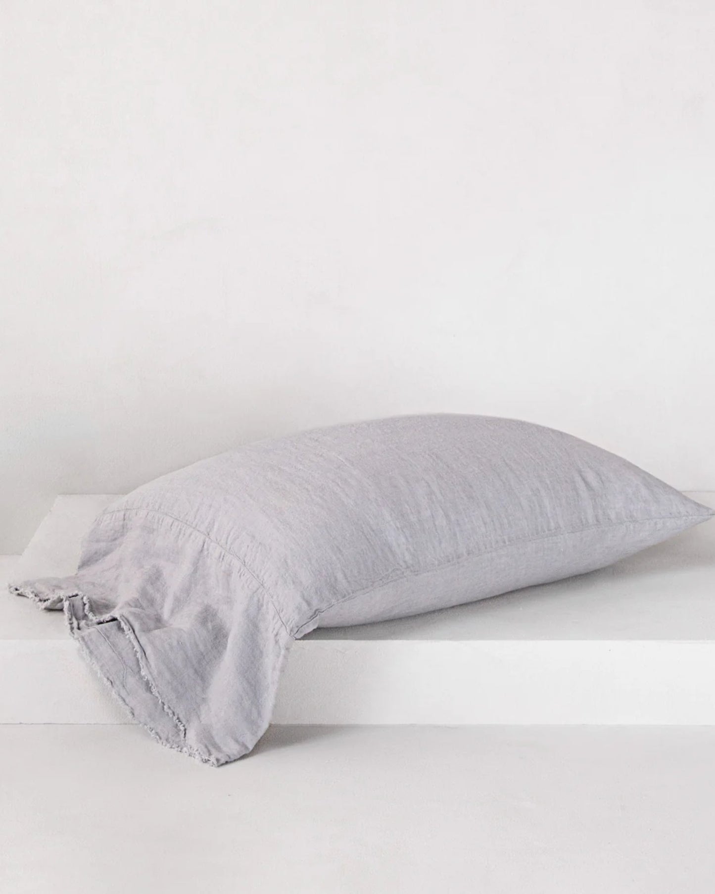 A HALE FLOCCA FLOPPY END PILLOWCASE by Hale Mercantile Co in light gray linen, featuring hand-tufted edges, rests on a white platform against a plain background. This pillowcase offers a soft, delicate texture and minimalist design that perfectly complements any pure linen bedding ensemble.