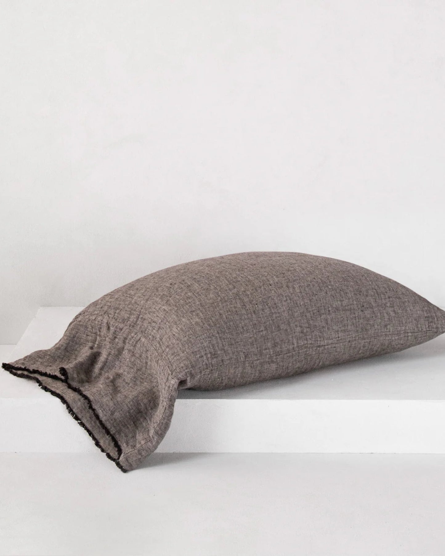 A HALE FLOCCA FLOPPY END PILLOWCASE by Hale Mercantile Co, featuring hand-tufted edges in brown, rests on a smooth, white surface. The backdrop is a plain white wall, enhancing the image's minimalist and clean appeal.