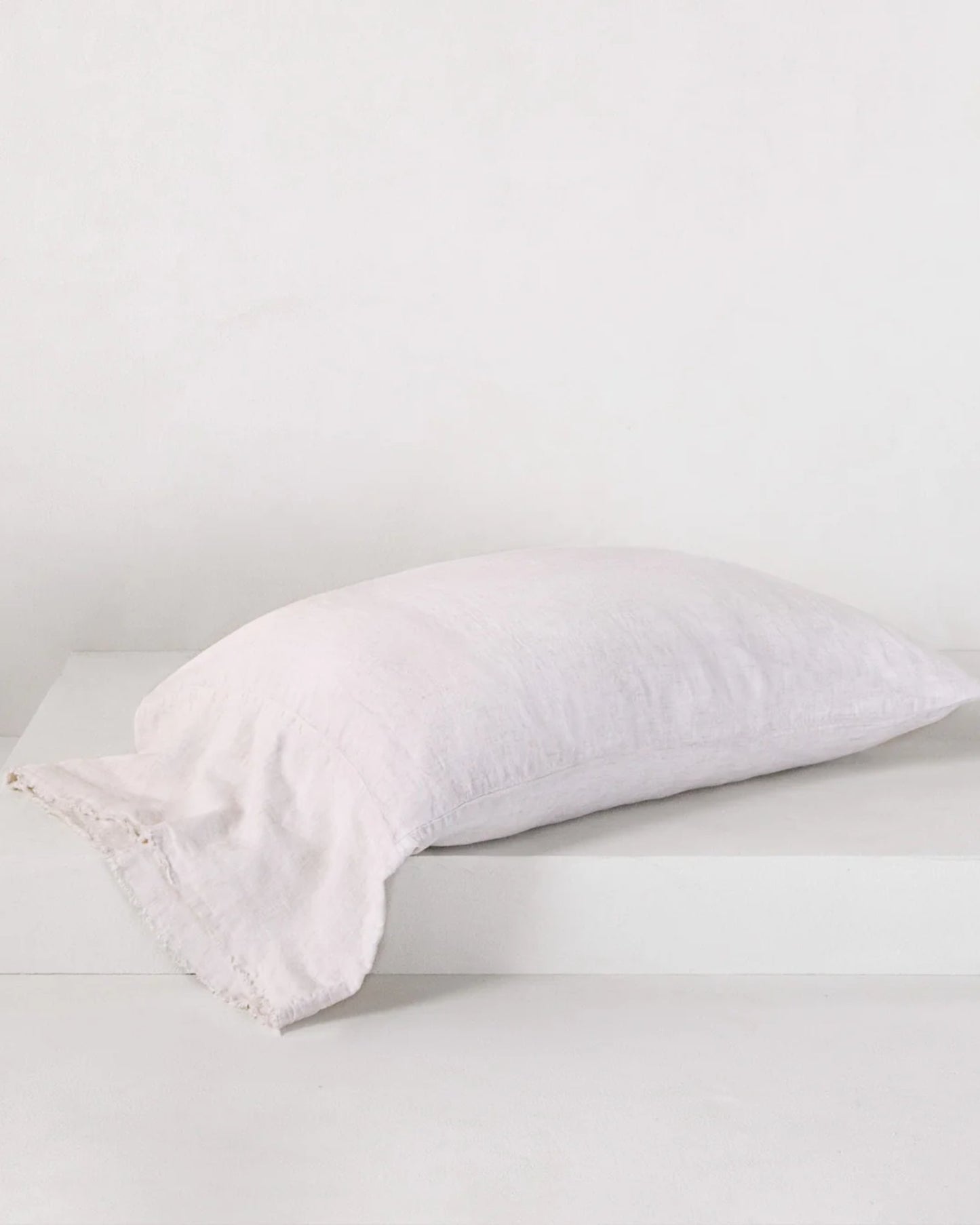 A soft, light pink pillow featuring the Hale Mercantile Co HALE FLOCCA FLOPPY END PILLOWCASE with hand-tufted edges rests on a white platform against a plain white background. The linen pillowcase enhances its simple and elegant design.