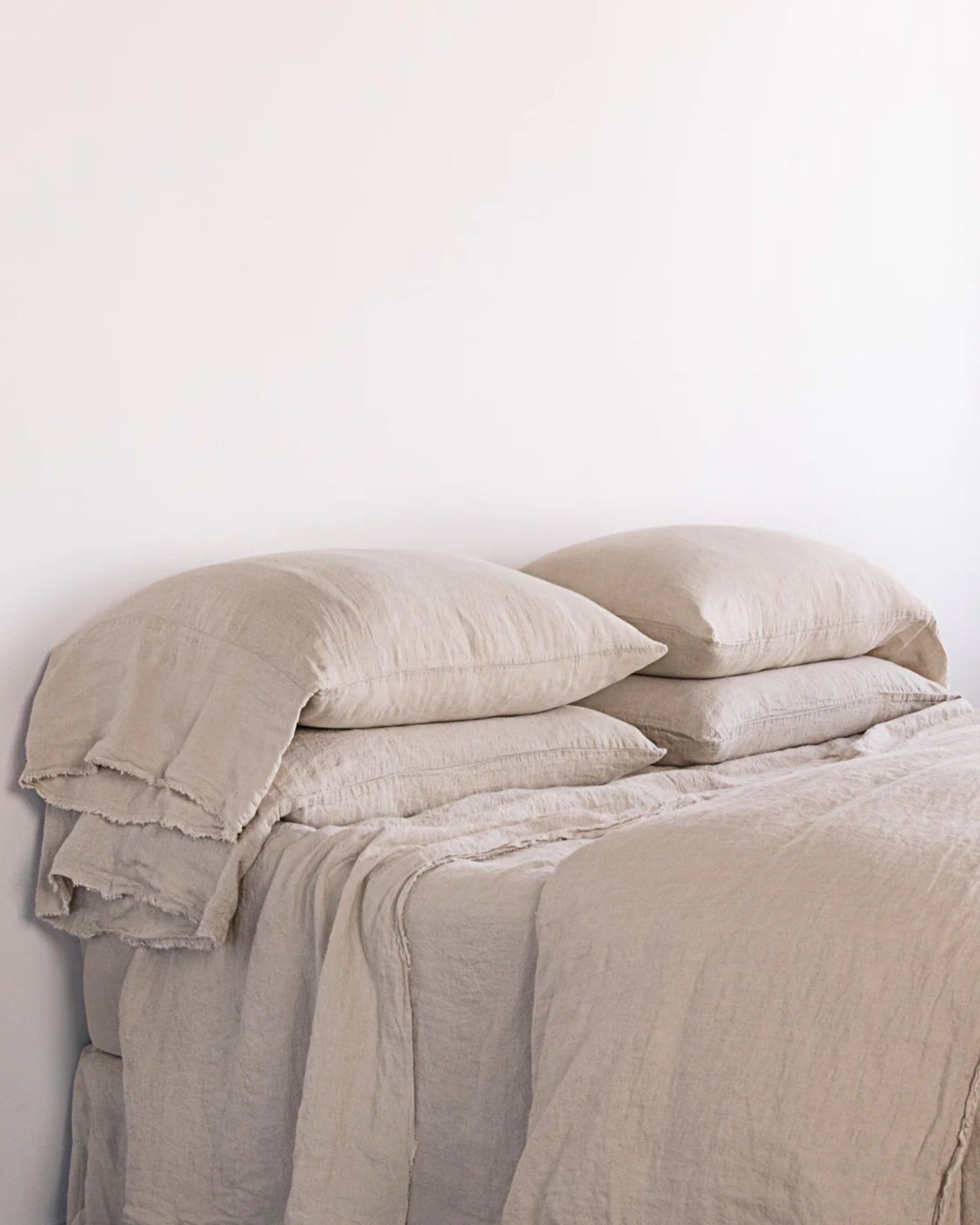 A neatly made bed with pure linen bedding and beige sheets against a plain white wall offers a minimalist and cozy appearance. The pillows, adorned with Hale Mercantile Co's Flocca Floppy End Pillowcases featuring hand-tufted edges, are stacked in pairs, and the bedding drapes softly over the mattress.