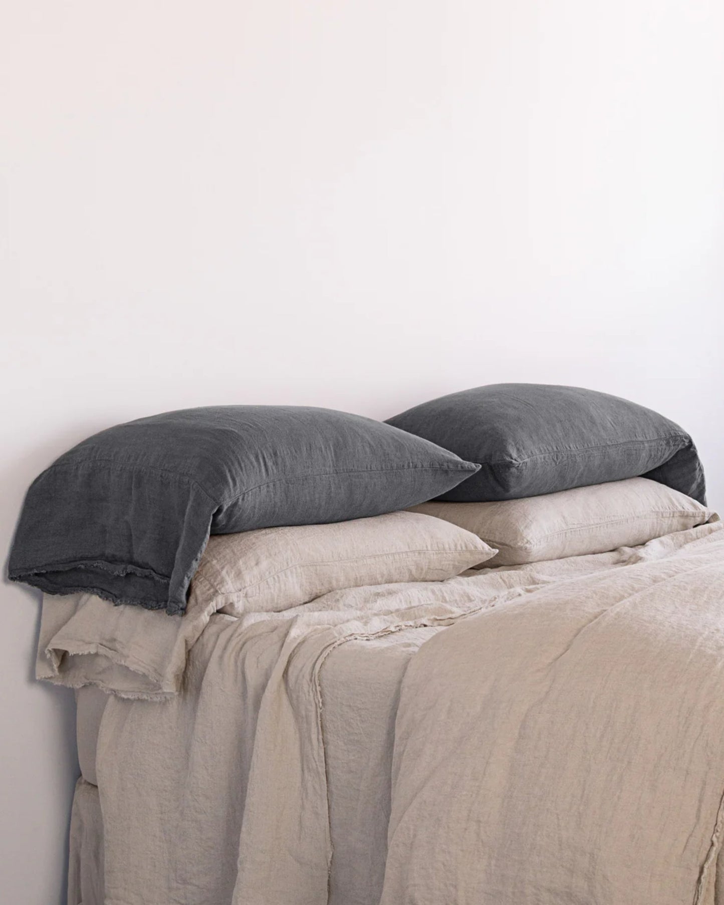 Against a plain white wall, a neatly made bed showcases two dark gray pillows and two beige ones, all encased in the HALE FLOCCA FLOPPY END PILLOWCASE by Hale Mercantile Co. The bed is enhanced by a beige, textured duvet crafted from pure linen bedding, offering a minimalist and cozy atmosphere.