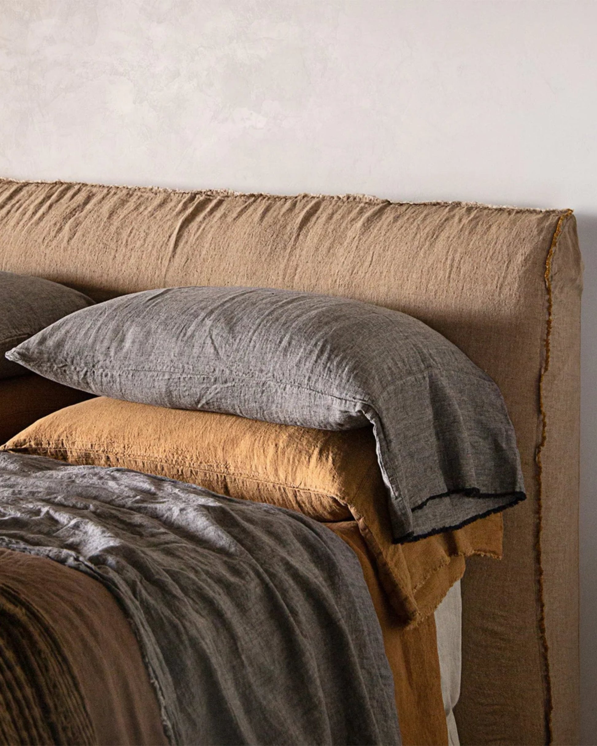 A neatly made bed showcases brown and gray textured linens with Flocca hand-tufted edges on the pure linen bedding set. The rustic, cozy appearance of the headboard, paired with the Hale Mercantile Co's HALE FLOCCA FLOPPY END PILLOWCASE, creates a warm and inviting atmosphere. The neutral wall behind perfectly complements the tones of the bedding.