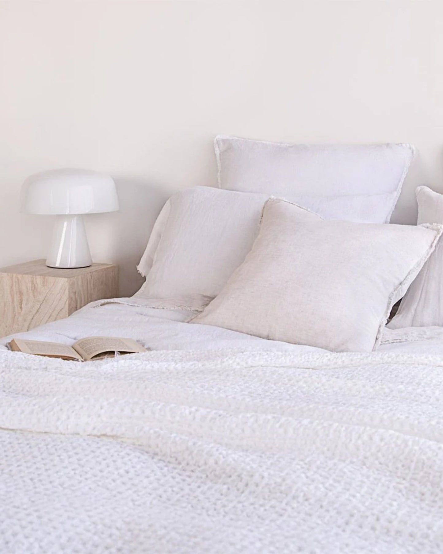 A cozy bedroom scene showcases a neatly made bed adorned with plush white pillows and the HALE FLOCCA THROW, a textured pure linen comforter by Hale Mercantile Co. An open book lies on the bed, while a wooden bedside table supports a sleek white lamp. The walls are painted in a soft, light hue.