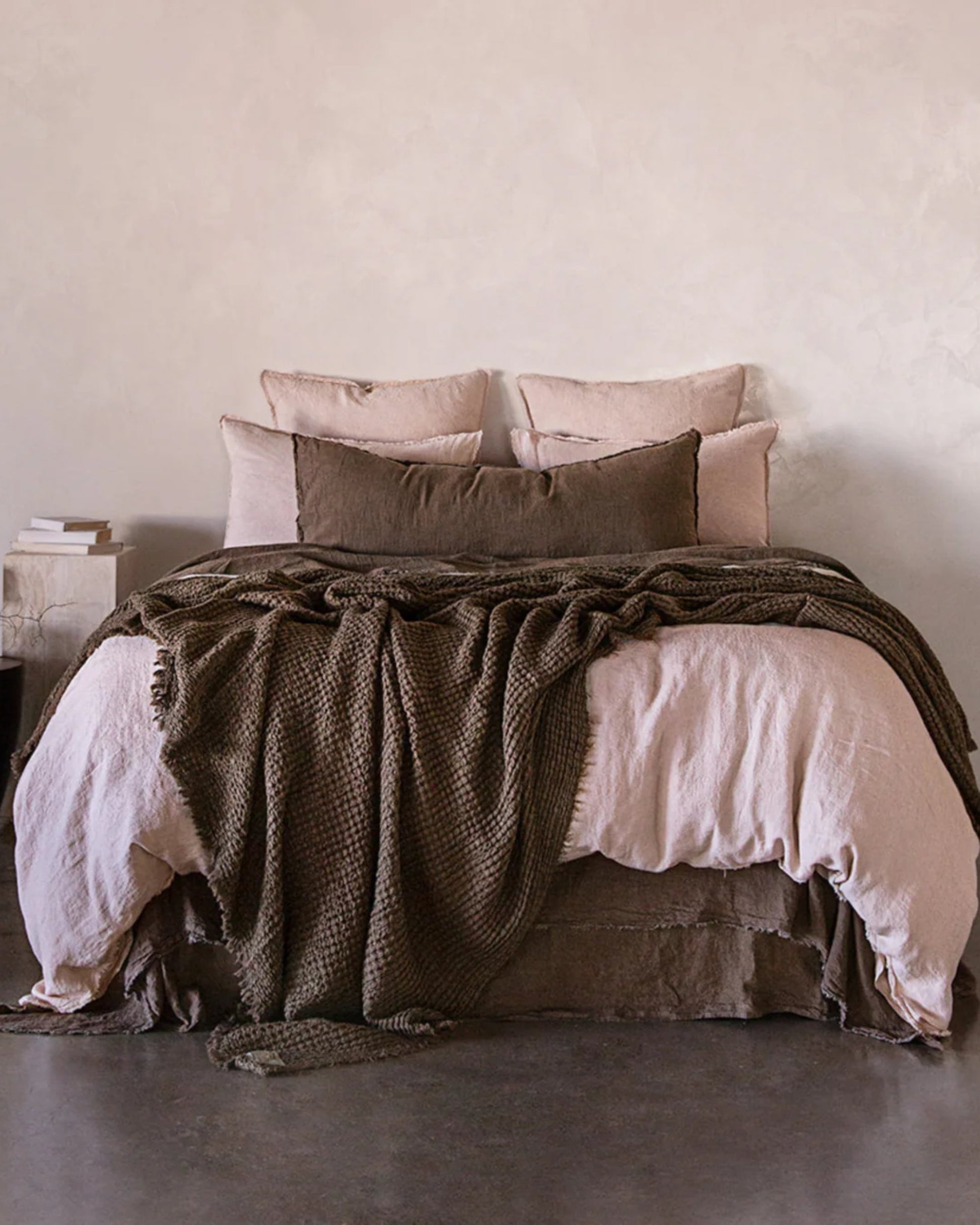 A cozy, well-appointed bed is adorned with light pink and brown pillows and the luxurious HALE FLOCCA THROW by Hale Mercantile Co. The bed is positioned against a neutral-colored wall, complemented by a small side table and books to the left. The polished concrete floor adds an elegant touch to the tranquil room.