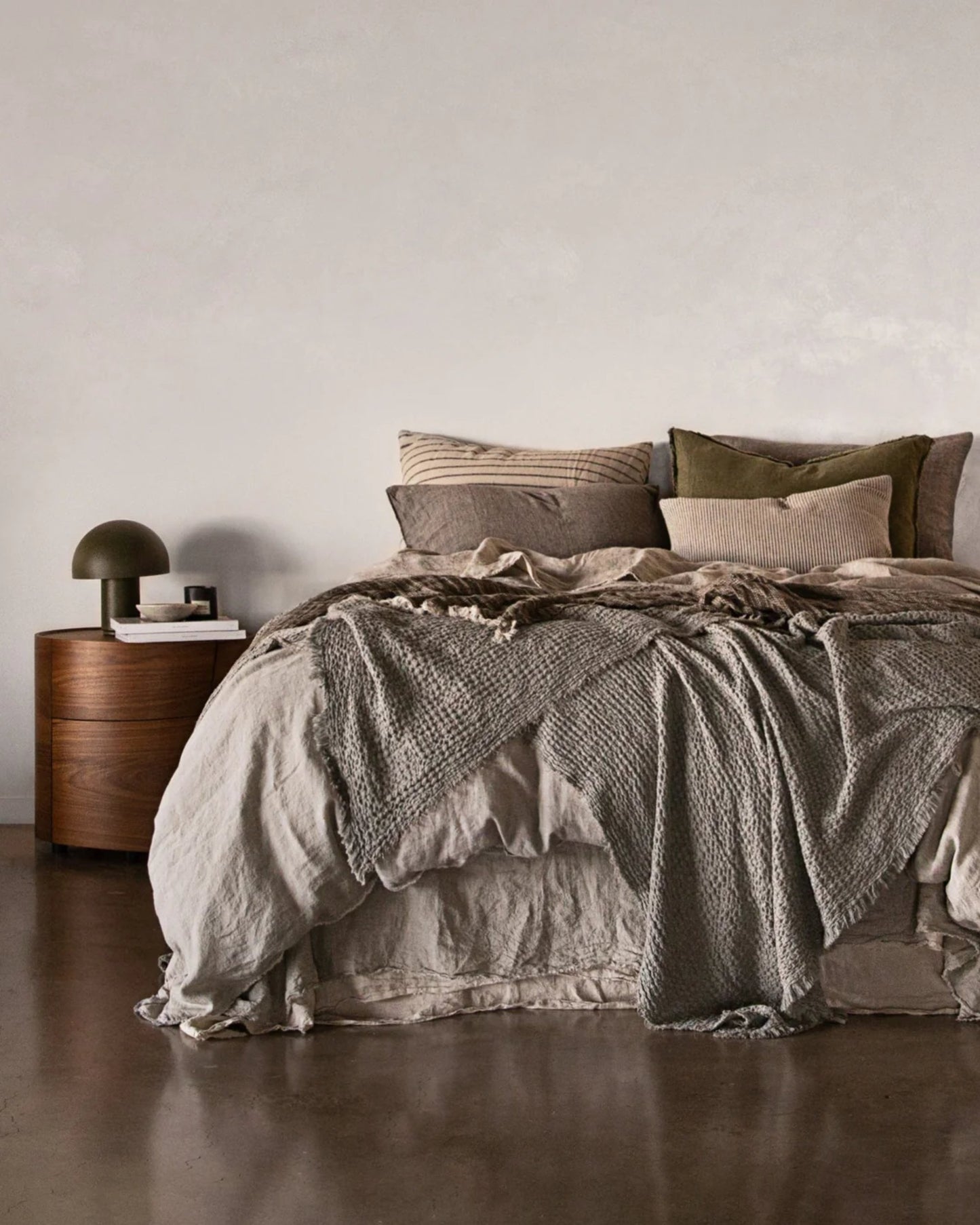 A cozy bedroom scene showcases a neatly made bed draped with the HALE FLOCCA THROW from Hale Mercantile Co, complemented by neutral-toned linens. Several pillows enhance comfort, while a round wooden nightstand supports a green lamp and stacked books, all set against a plain, light-colored wall.