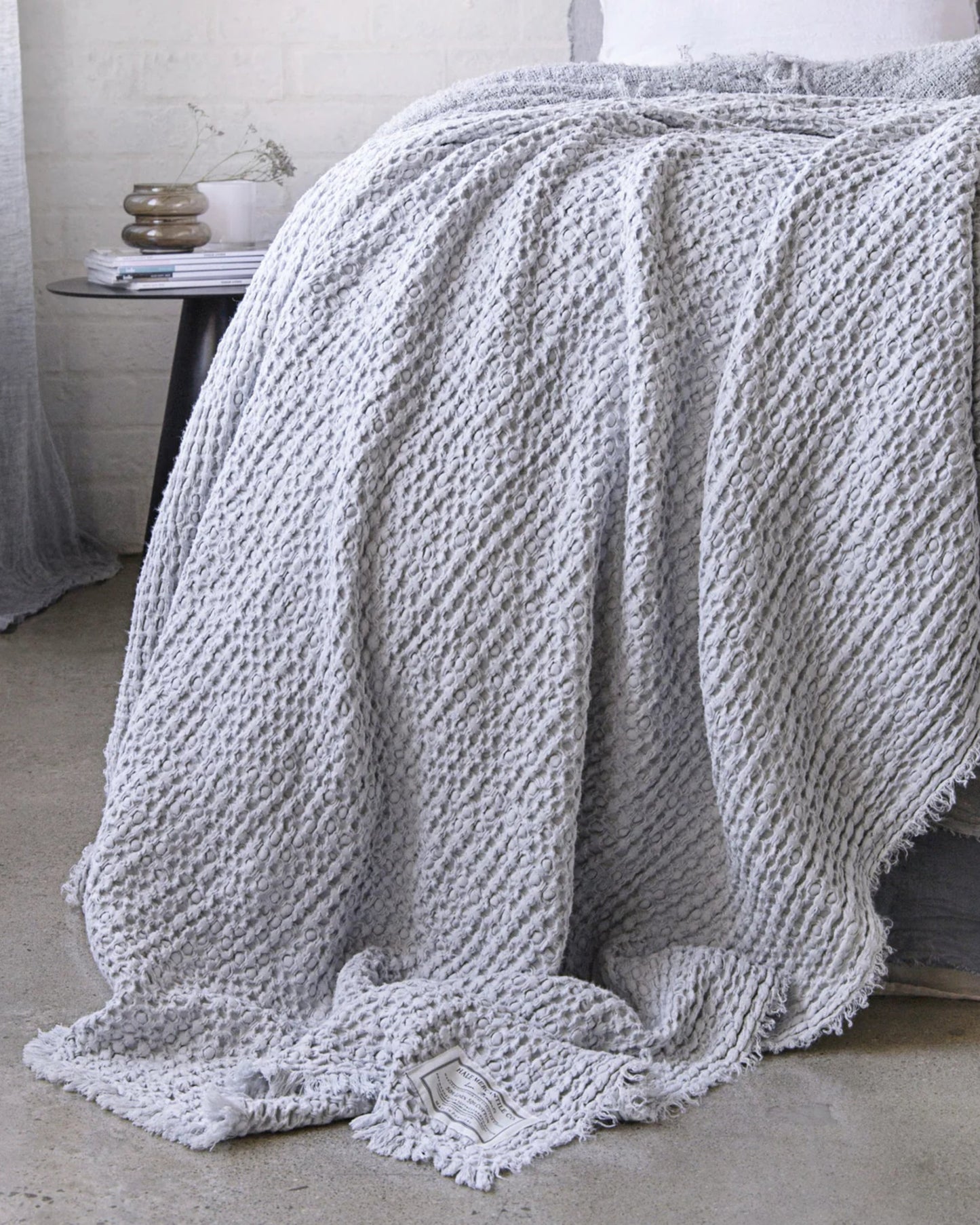 The HALE FLOCCA THROW by Hale Mercantile Co, featuring a cozy textured gray linen with fringe edges, is elegantly draped over the bed. The light concrete floor and a small table adorned with stacked books and a plant contribute to the serene, minimalist ambiance of the space, enhancing its luxurious aura.