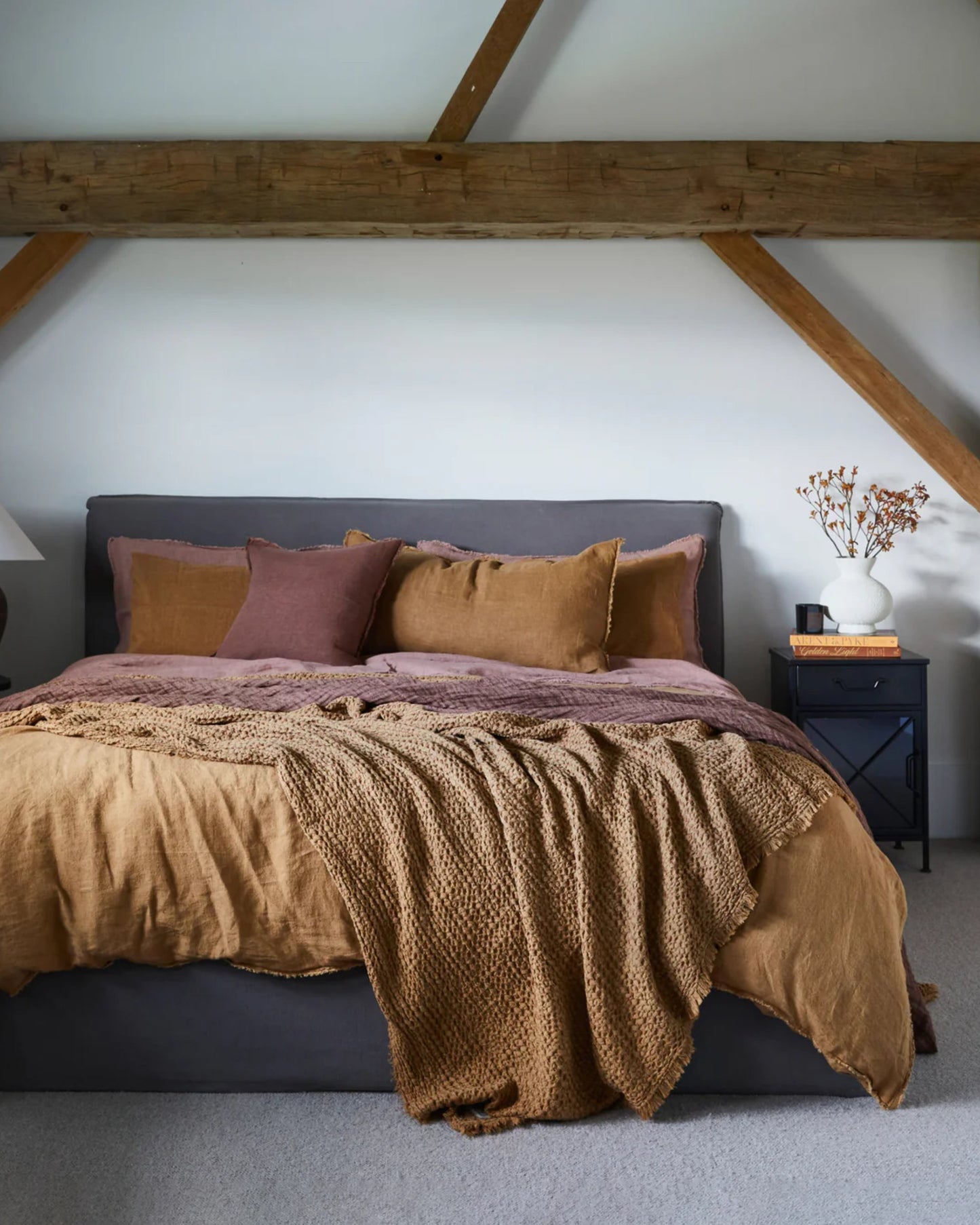 In a cozy bedroom, you'll find a large bed with an elegant gray frame adorned with earth-toned bedding, including the luxurious HALE FLOCCA THROW by Hale Mercantile Co in mustard and mauve linen. Positioned next to the bed is a sleek black nightstand, which holds both a plant and a book. The white walls are accented by visible wooden beams.